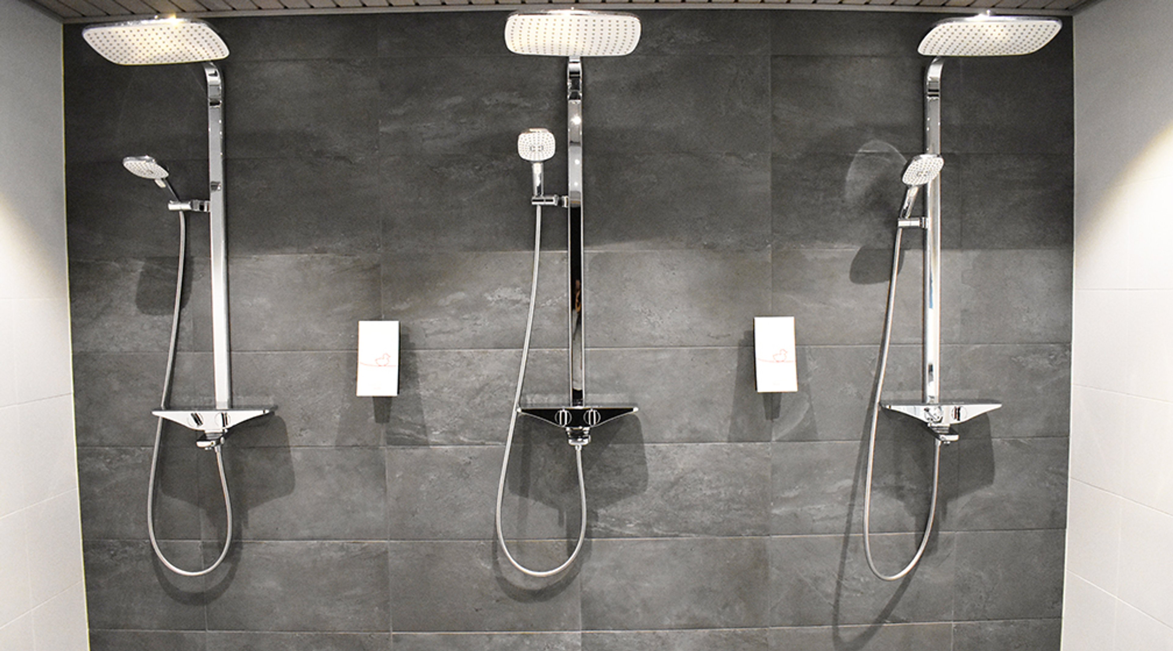 Moderneja suihkuja kolme riviä harmaan laatoitetussa kylpyhuoneessa, jokaisessa kiiltävä suihkupää ja käsisuihku telineessä.