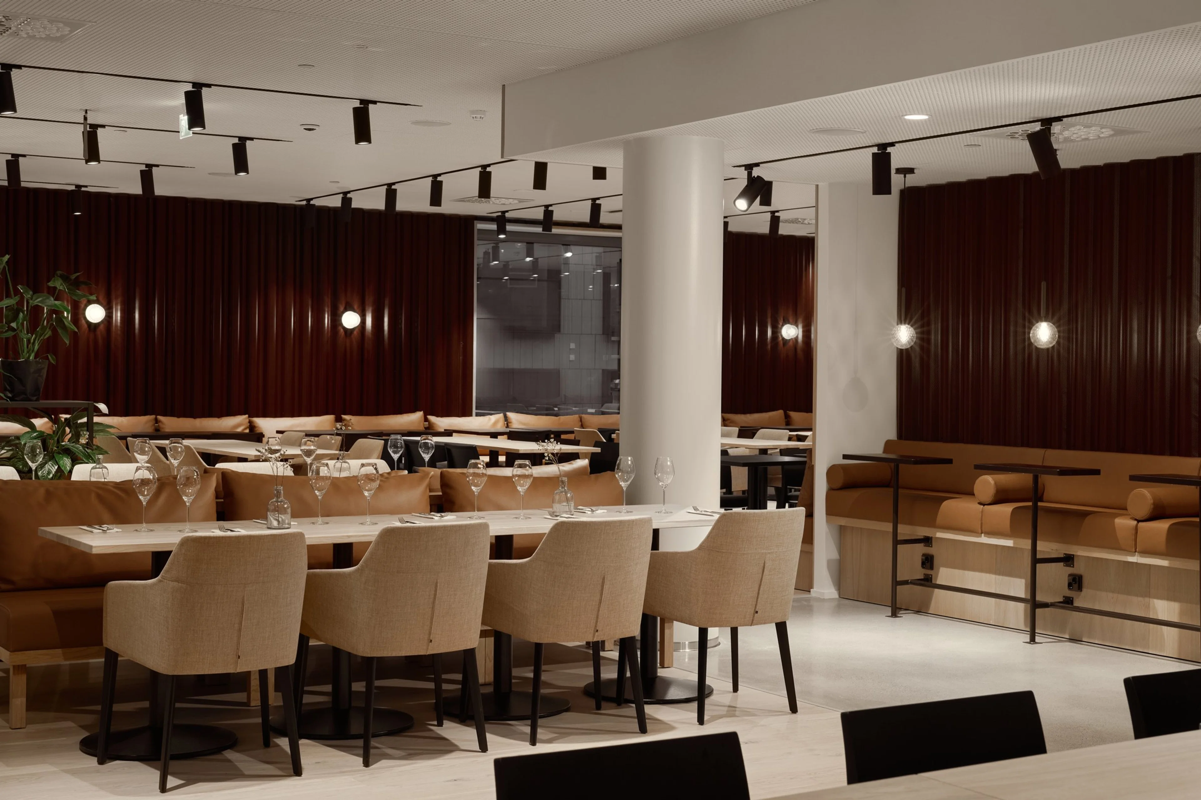 Moderni ravintola ruskein paneliseinin ja beigein istuimin. Avoin tila, jossa pöytiä ja tuoleja tarjoiluvalmiina. Valaistus spot-valoilla ja seinävalaisimilla.