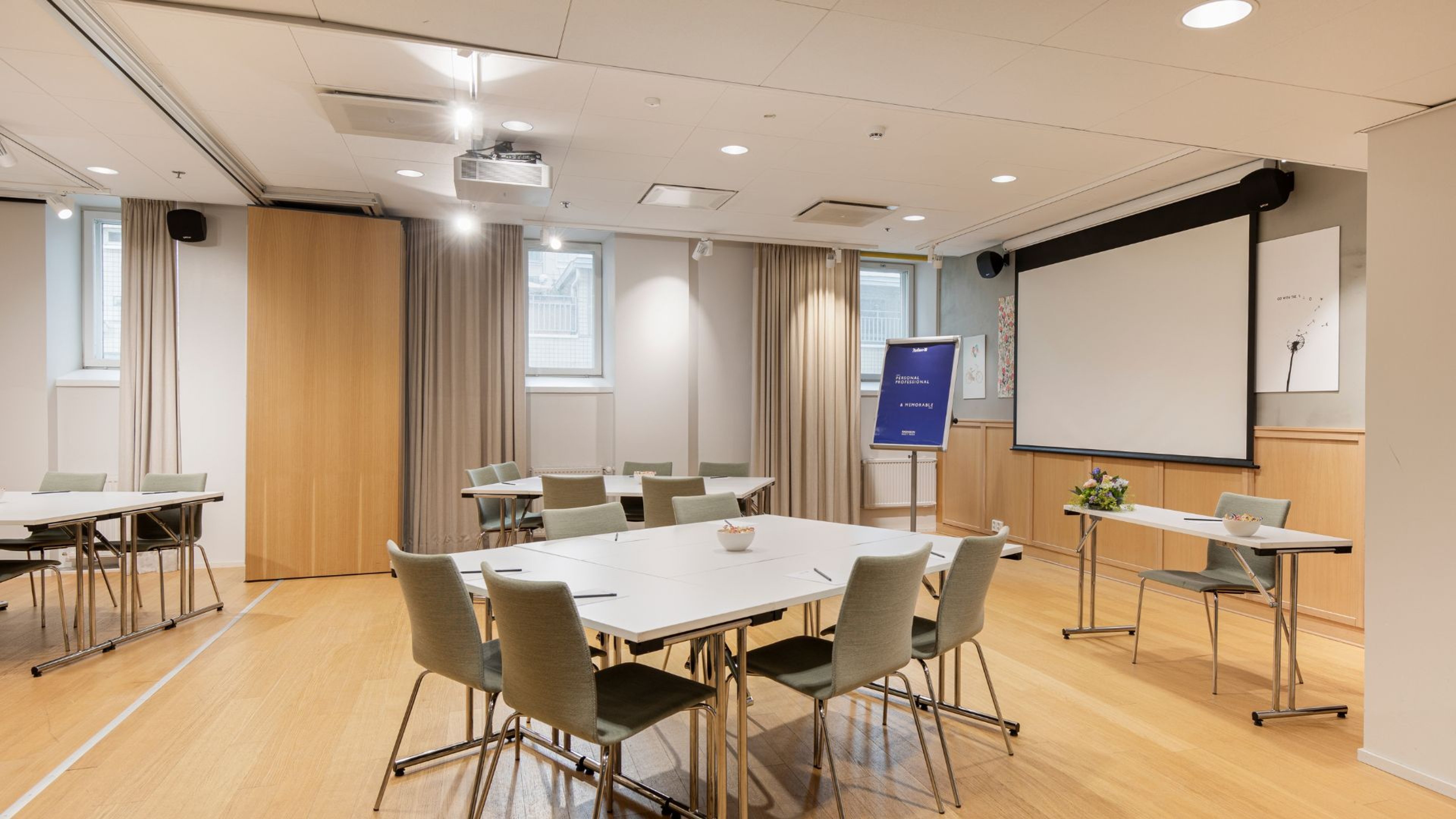 Valoisa kokoustila puulattialla ja liikuteltavilla pöydillä. Edessä valkokangas ja digitaalinen näyttö, luonnonvaloa ikkunoista.