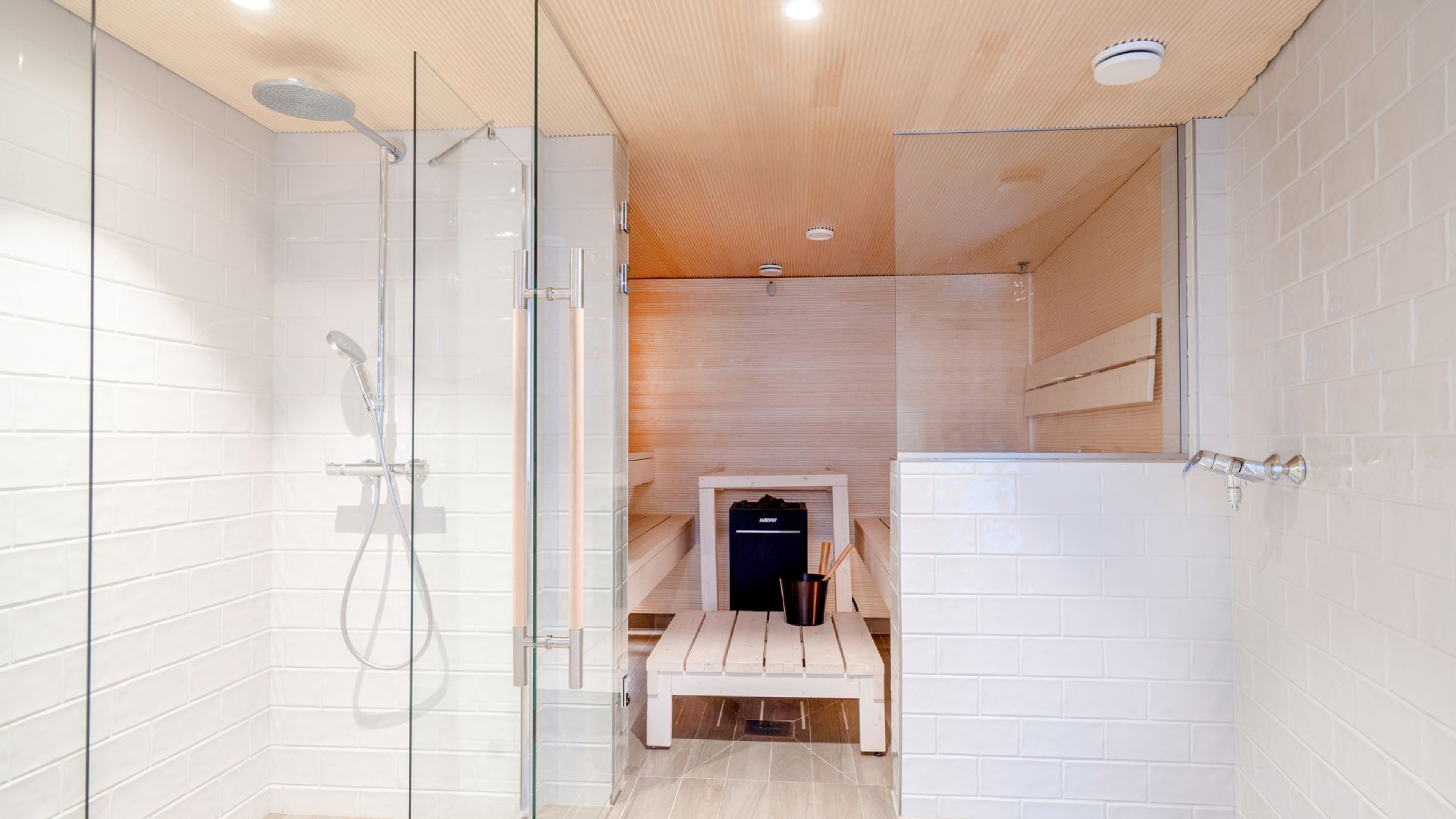 Moderni saunaosasto lasiovisella suihkulla ja vaaleasävyisellä puupaneloinnilla. Saunassa valkoinen puinen istuin ja kiuas.