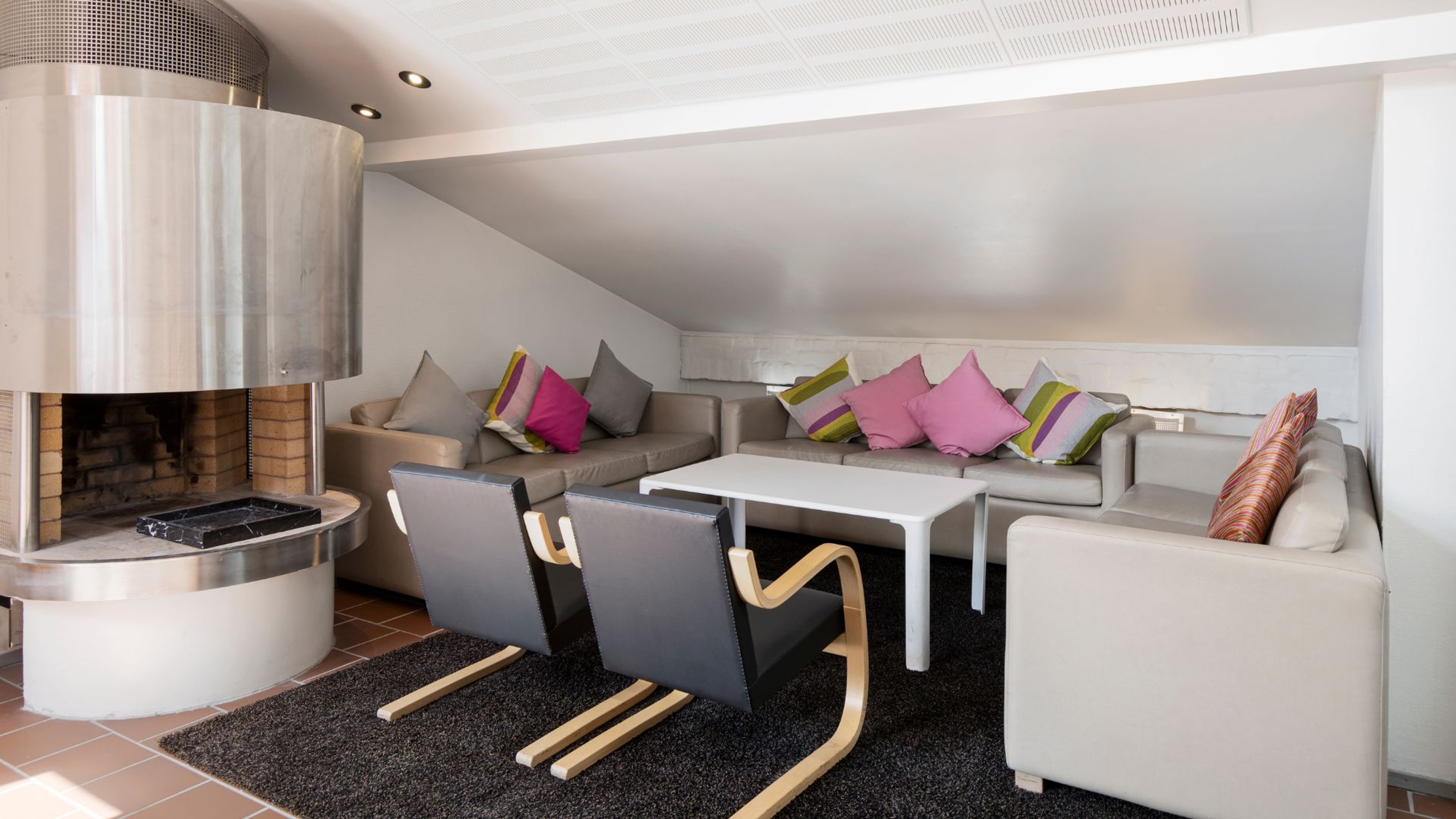 Moderni olohuone, jossa vaaleat sohvat, värikkäitä tyynyjä, kaksi mustaa nojatuolia puisilla jaloilla ja metallinen takka. Lattialla tumma matto.