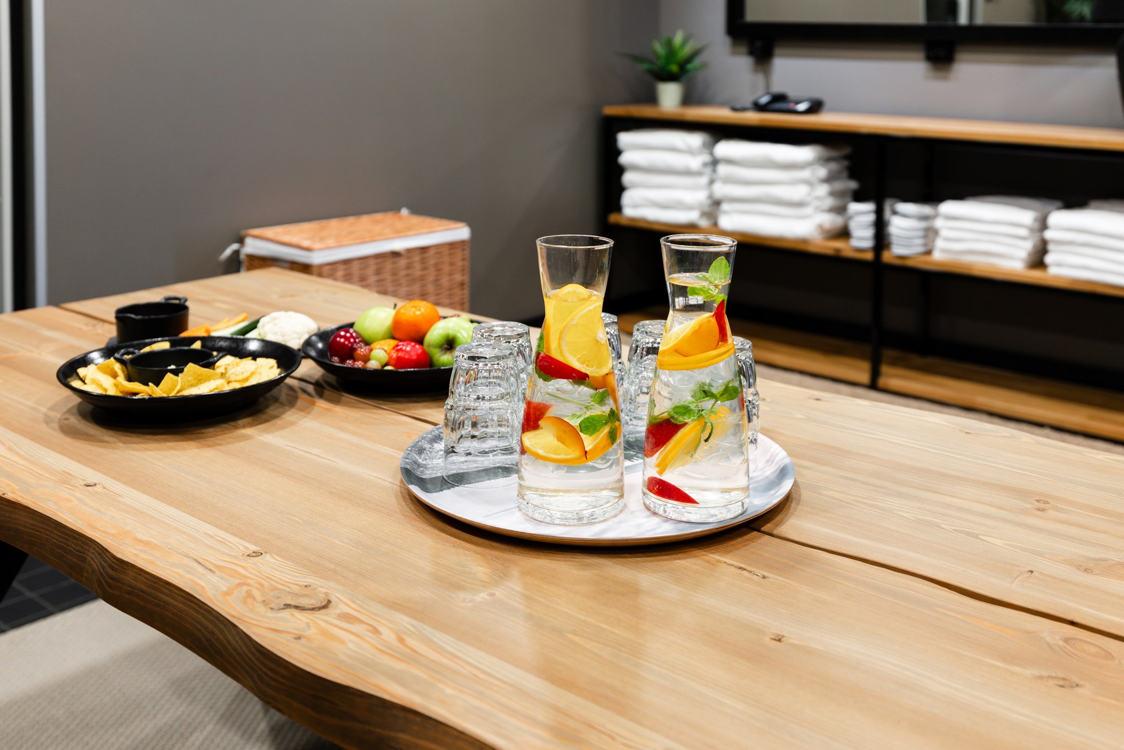 Bright relaxation room with wooden table featuring fruit-infused water in carafes and healthy snacks. Soft floor surface and white towels on shelving.