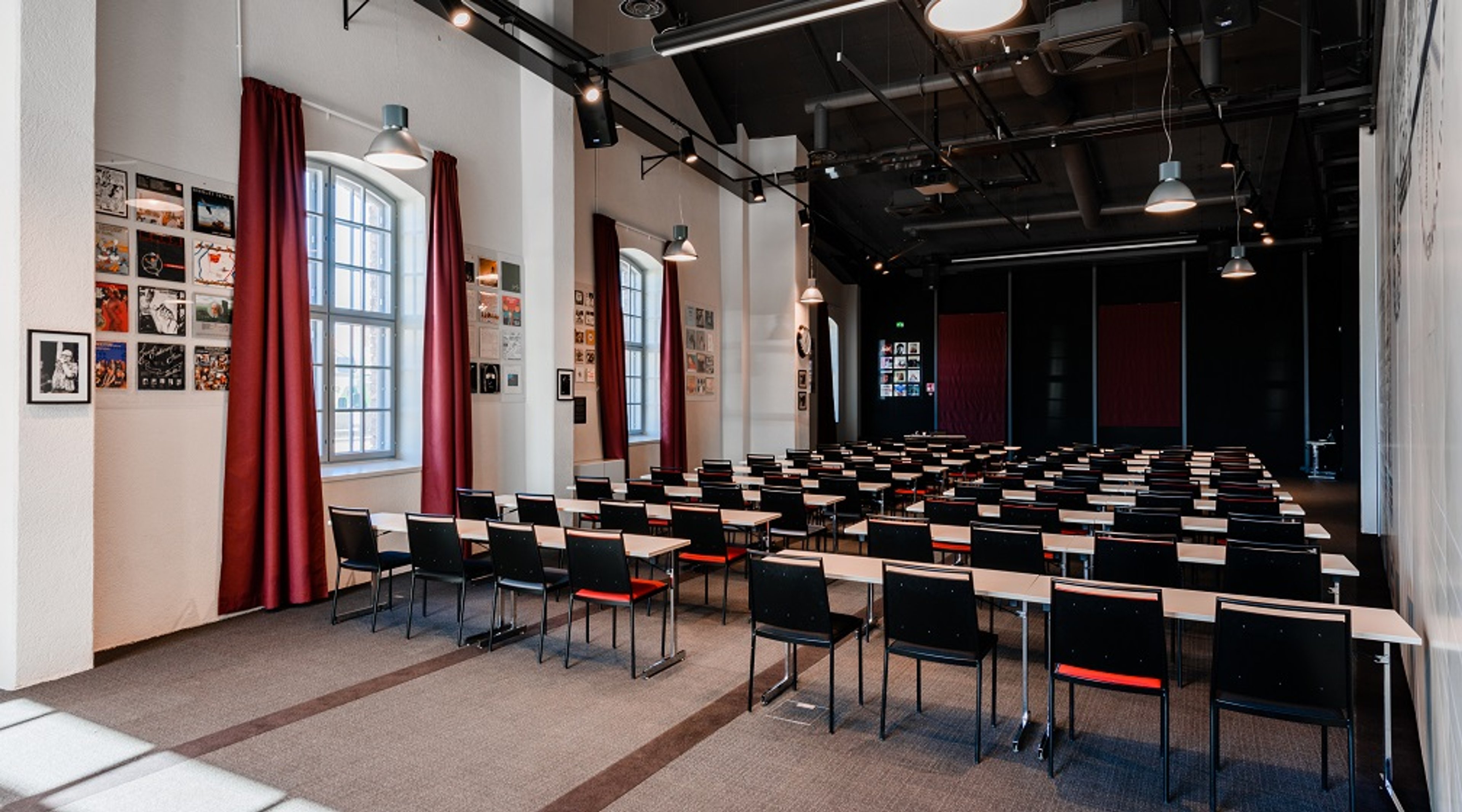 Auditoriotila, jossa on useita rivejä tuoleja, punaisia verhoja ikkunoiden edessä, korkeakatto valaisimilla ja valokuvia seinillä.