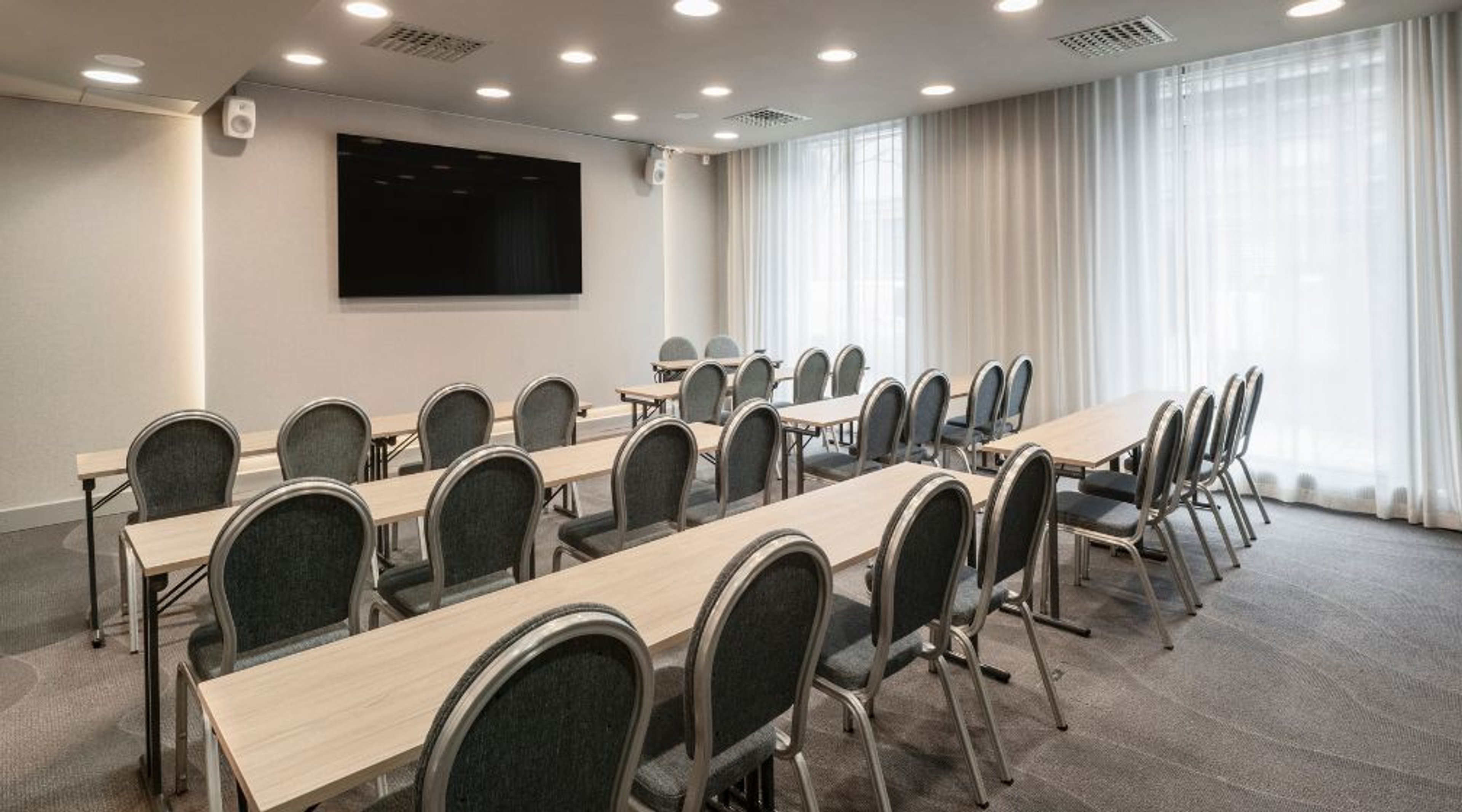 Valoisa kokoushuone, jossa on pitkät pöydät tuoleineen luokkamuodostelmassa, suuret ikkunat verhoineen ja seinällä musta näyttö.