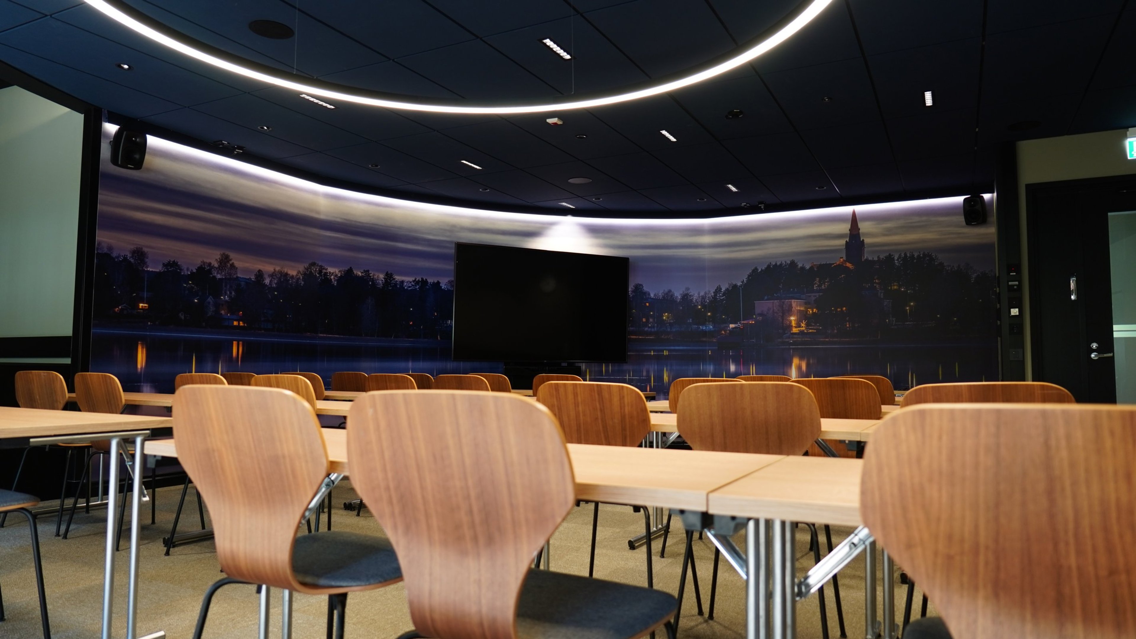 Kokoustila, jossa puiset tuolit ja pöydät. Seinällä panoraamanäyttö iltamaisemasta järven ja kirkon kanssa. Huoneessa tasainen lattia ja kaarevat valot katossa.