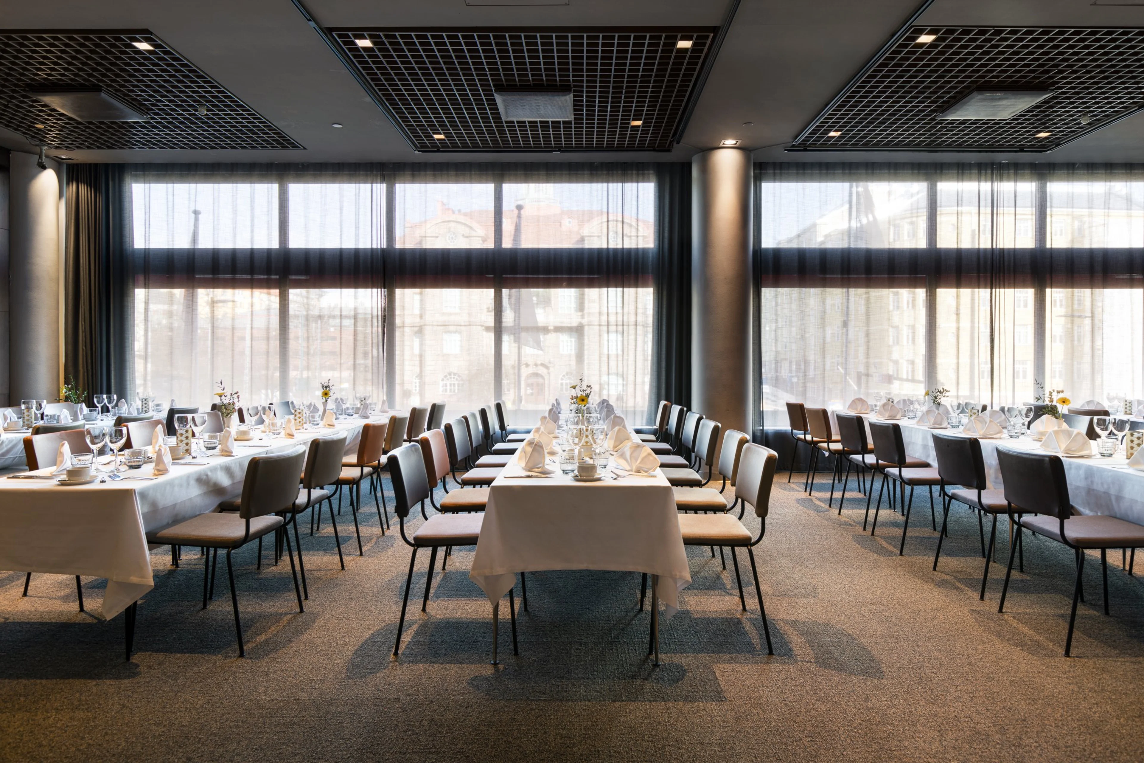 Moderni ravintolasali, jossa on valkoisilla liinoilla katetut pöydät, tyylikkäät tuolit ja suuret ikkunat. Tasainen lattiapinta ja leveät kulkuväylät pöytien välissä.