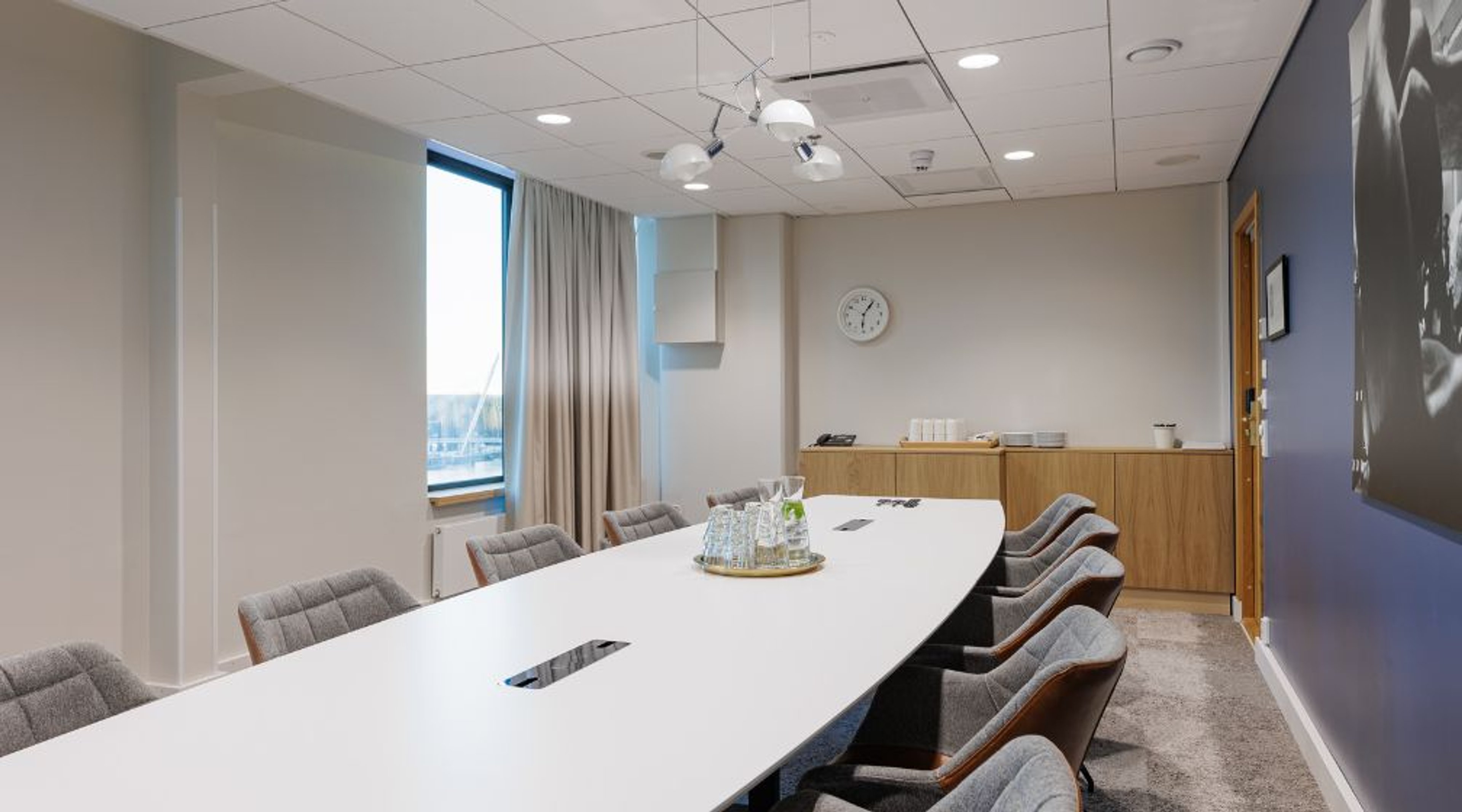 Moderni neuvottelutila, jossa pitkä valkoinen pöytä, harmaat tuolit, ikkunanäkymä ja puinen tarjoilukaappi. Tilassa kova lattiamateriaali ja hyvä valaistus.