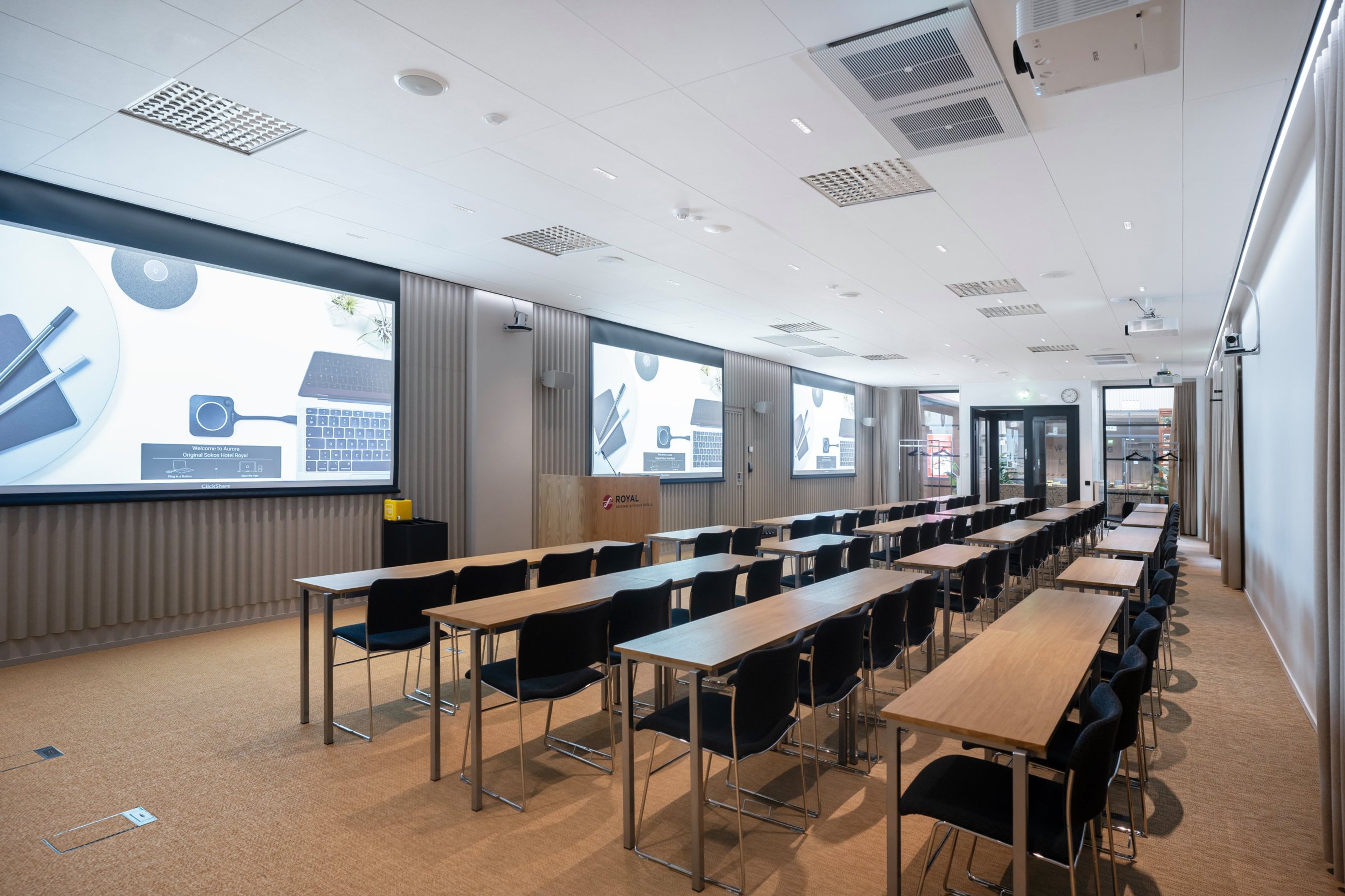 Moderni luokkahuone pitkillä pöydillä ja tuoleilla, suuret valkokankaat seinillä, tasainen lattiapinta ja hyvä valaistus.