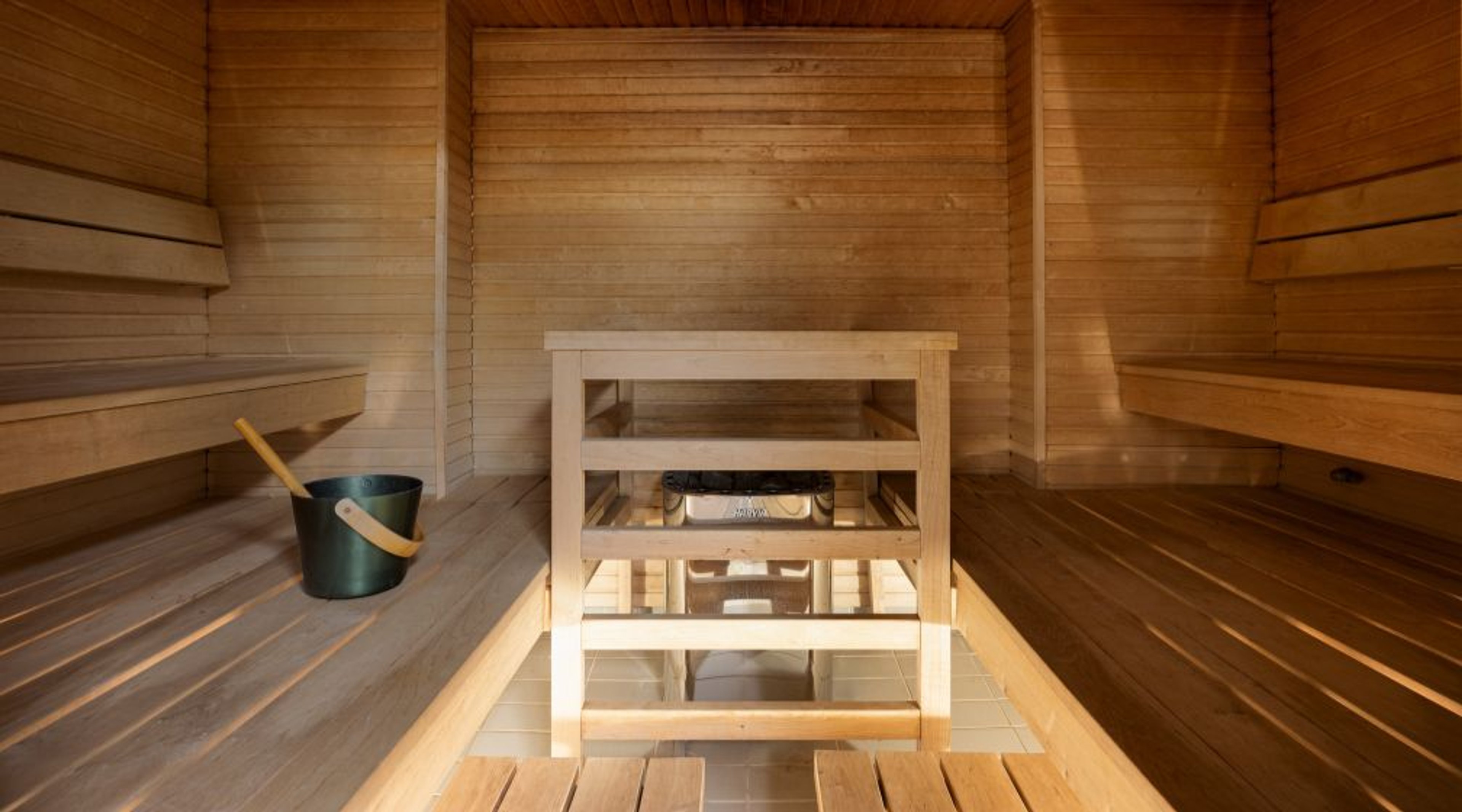 Sauna, jossa portaittaiset penkit ja puukiuas keskellä lattiaa. Vihta ja ämpäri vasemmalla penkillä.