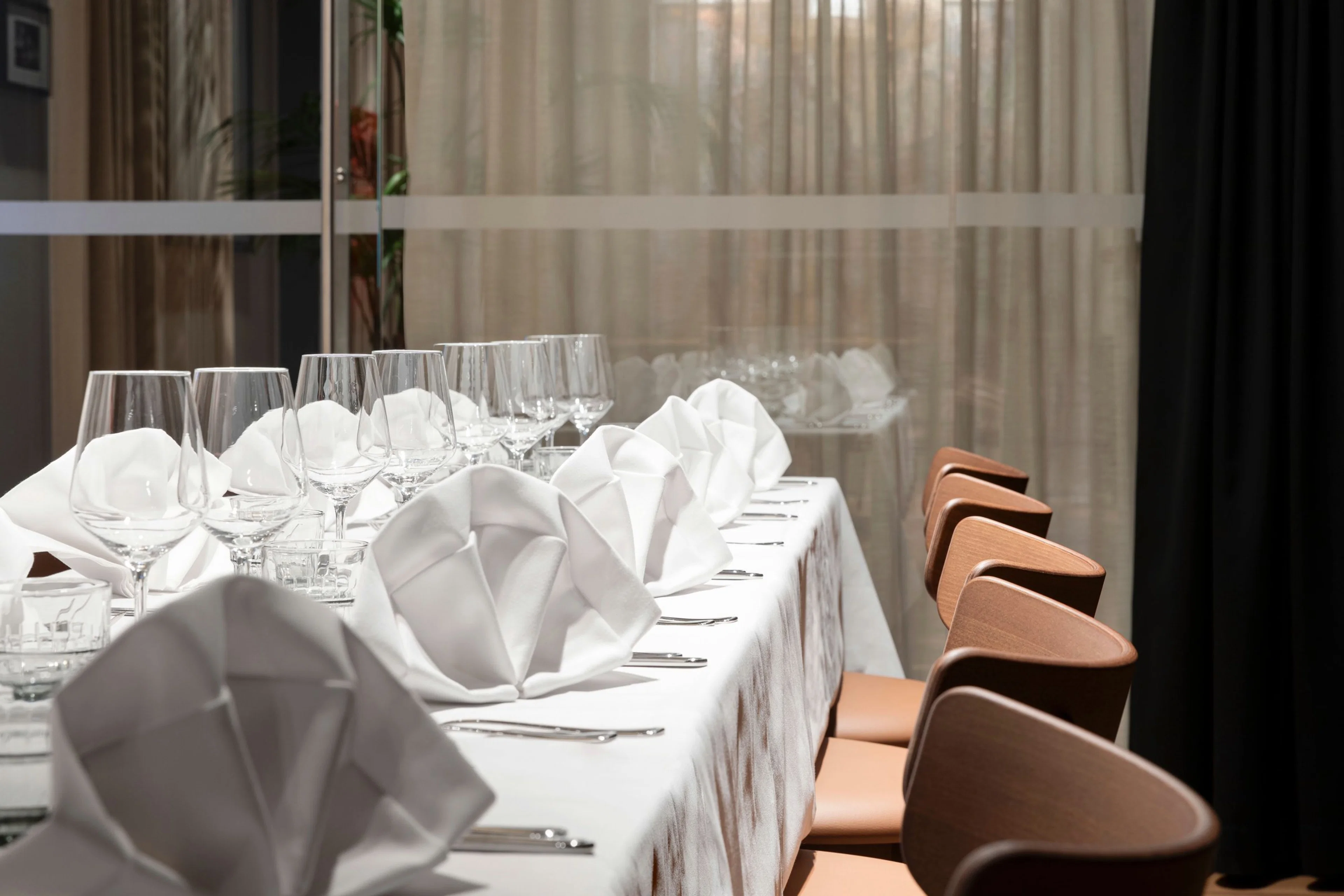 Elegantisti katettu pöytä valkoisella liinalla, taitellut lautasliinat, viinilasit ja puiset tuolit. Taustalla läpikuultava verho.
