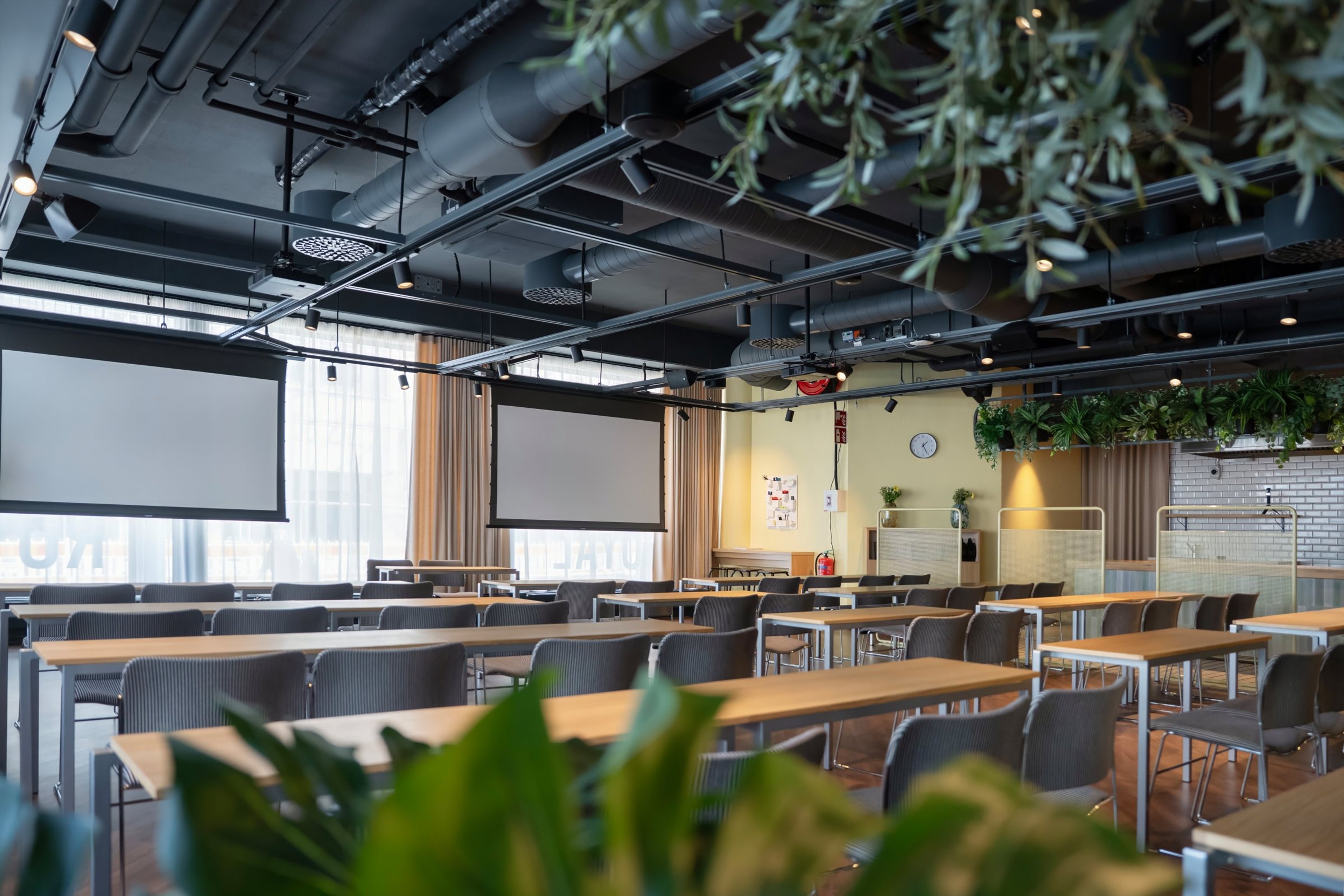 Moderni kokoustila puisilla pöydillä ja harmailla tuoleilla. Tilassa on kaksi projektorikangasta, viherkasveja ja teollinen katto.