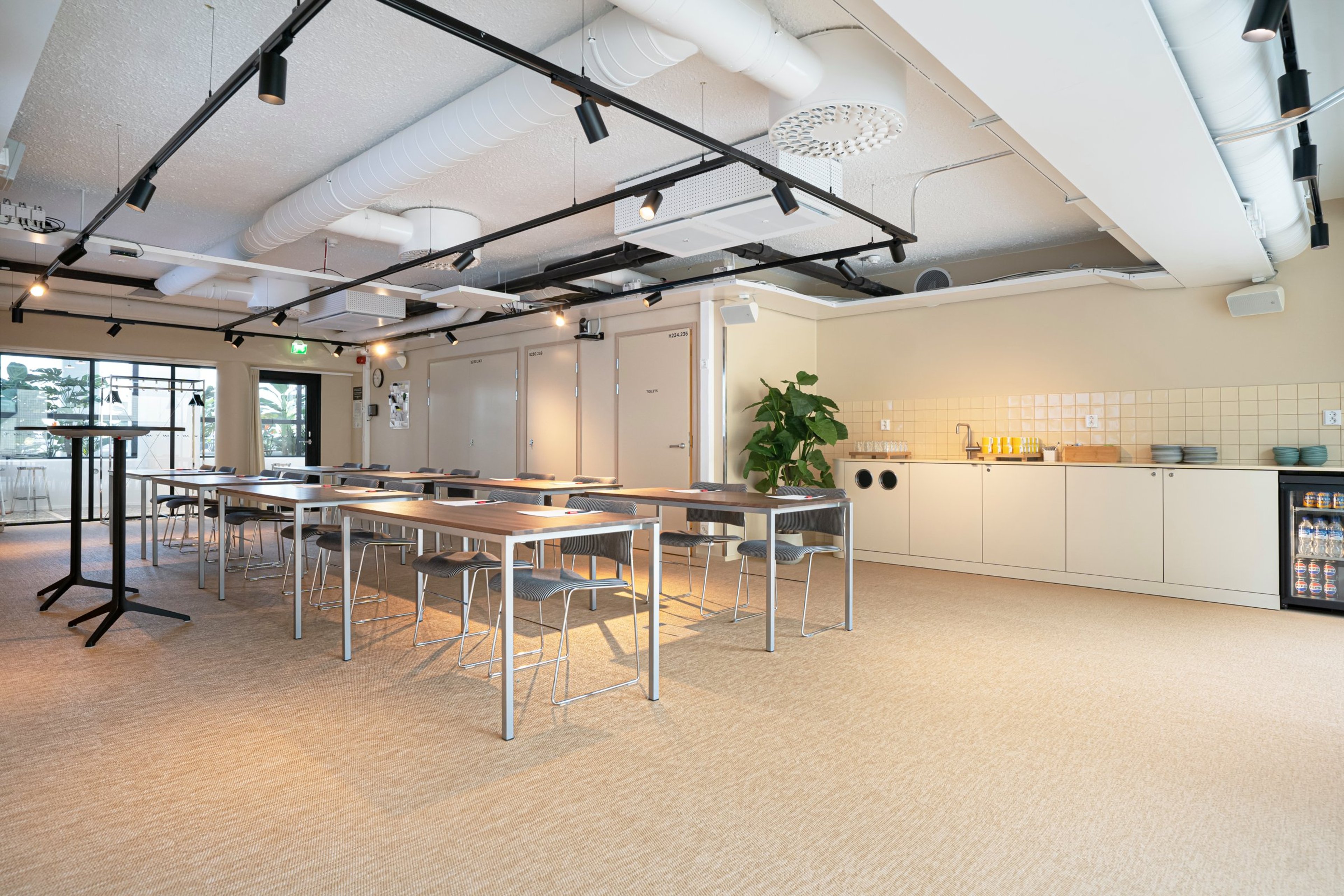 Moderni kokoushuone, jossa pitkiä pöytiä ja tuoleja, pieni keittiönurkkaus ja tasainen lattia. Iso ikkunaseinä tuo luonnonvaloa tilaan.