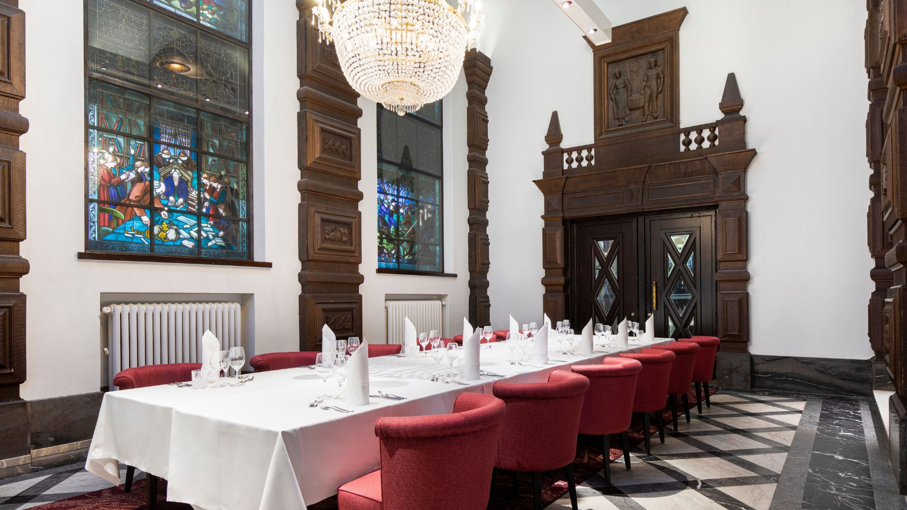 Juhlatila, jossa pitkä pöytä punaisilla tuoleilla. Tilassa koristeelliset lasimaalausikkunat, kristallikruunu ja puiset koristeelliset seinäkoristeet. Mustavalkoinen marmorimainen lattia.