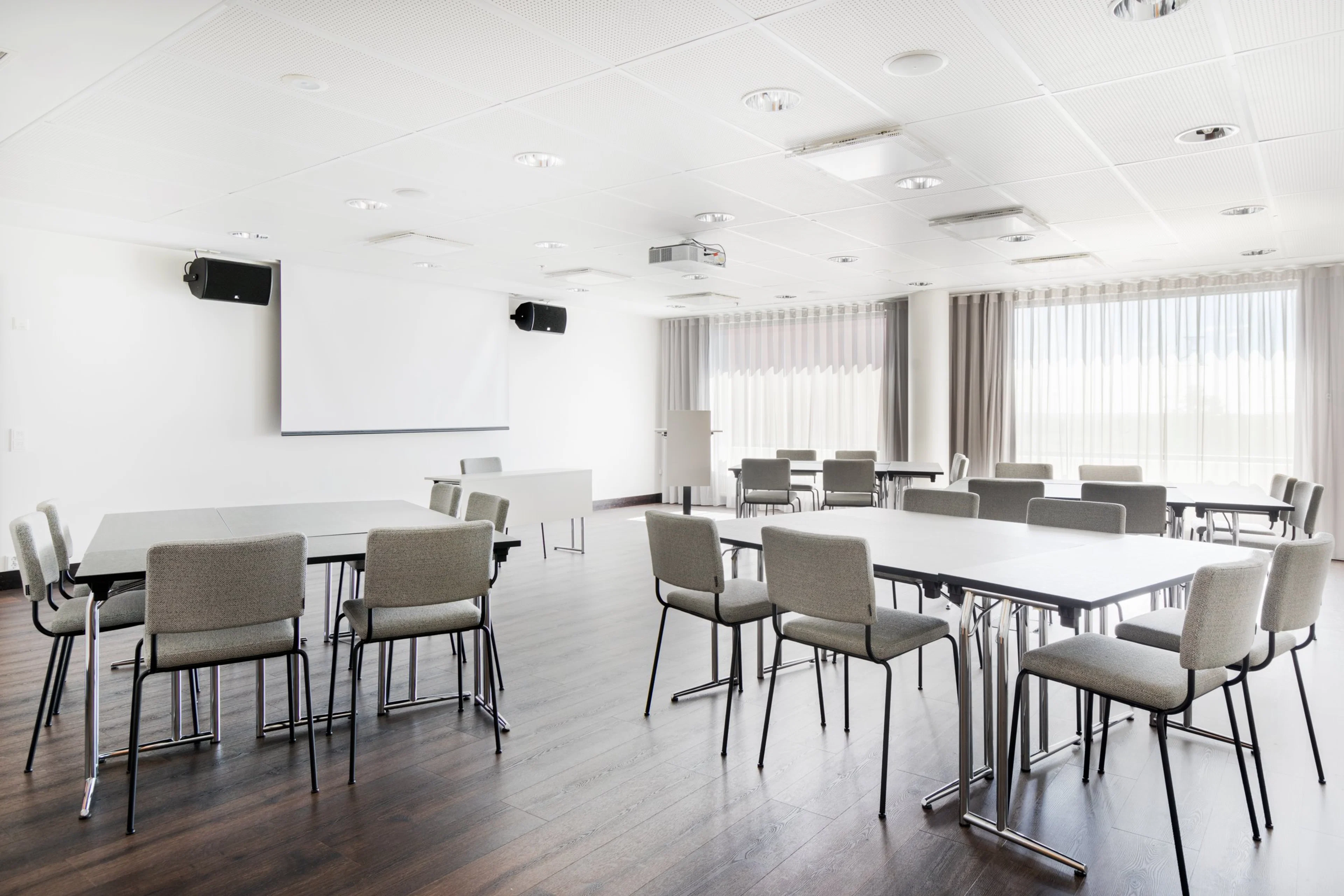 Moderni kokoustila valkoisilla pöydillä ja vaaleilla tuoleilla. Huoneessa on puinen lattia, projektori ja verhot suurissa ikkunoissa.