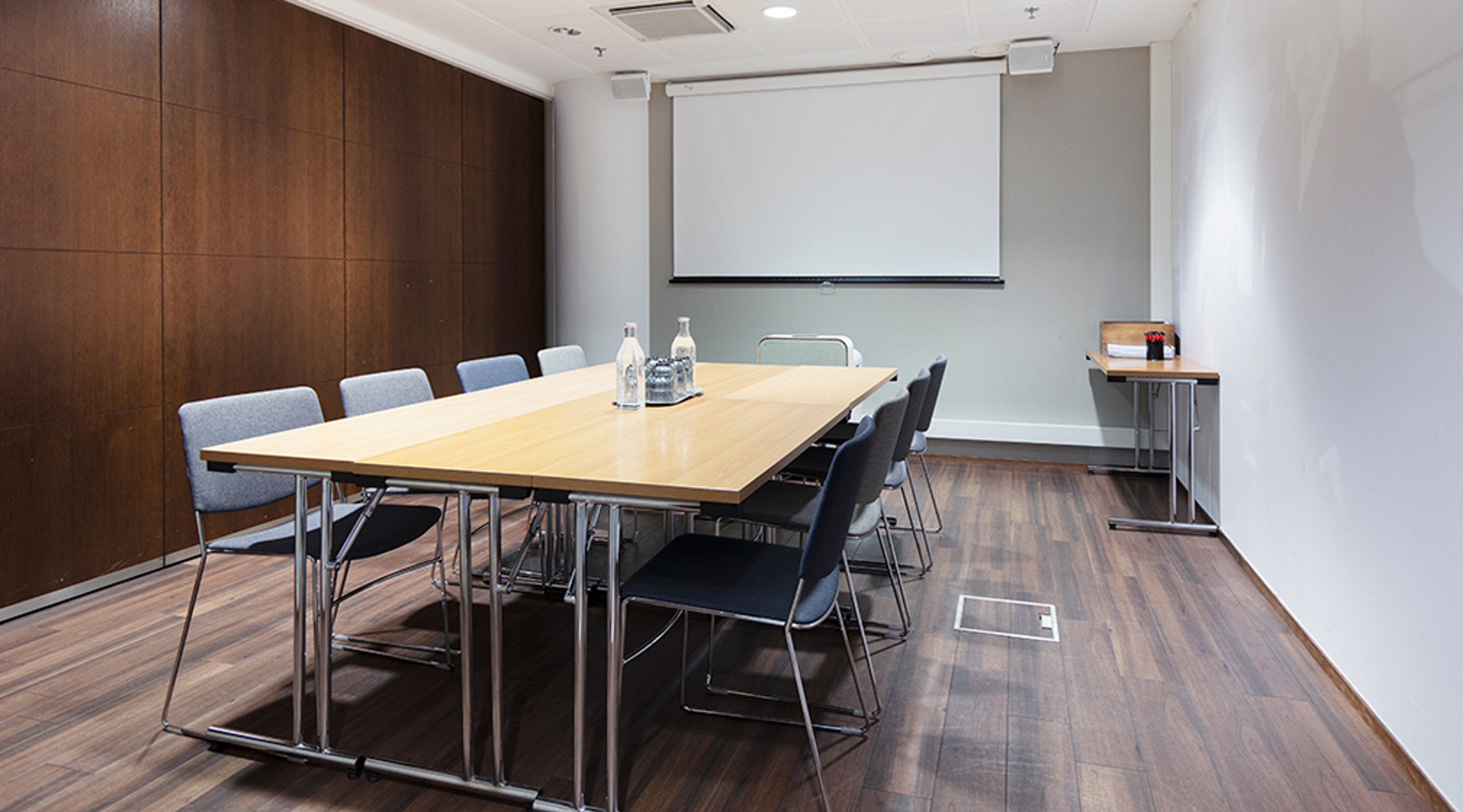 Moderni neuvotteluhuone puisella pöydällä, harmailla tuoleilla ja valkoisella esityskankaalla. Ruskea puupaneeli seinällä ja puulattia.