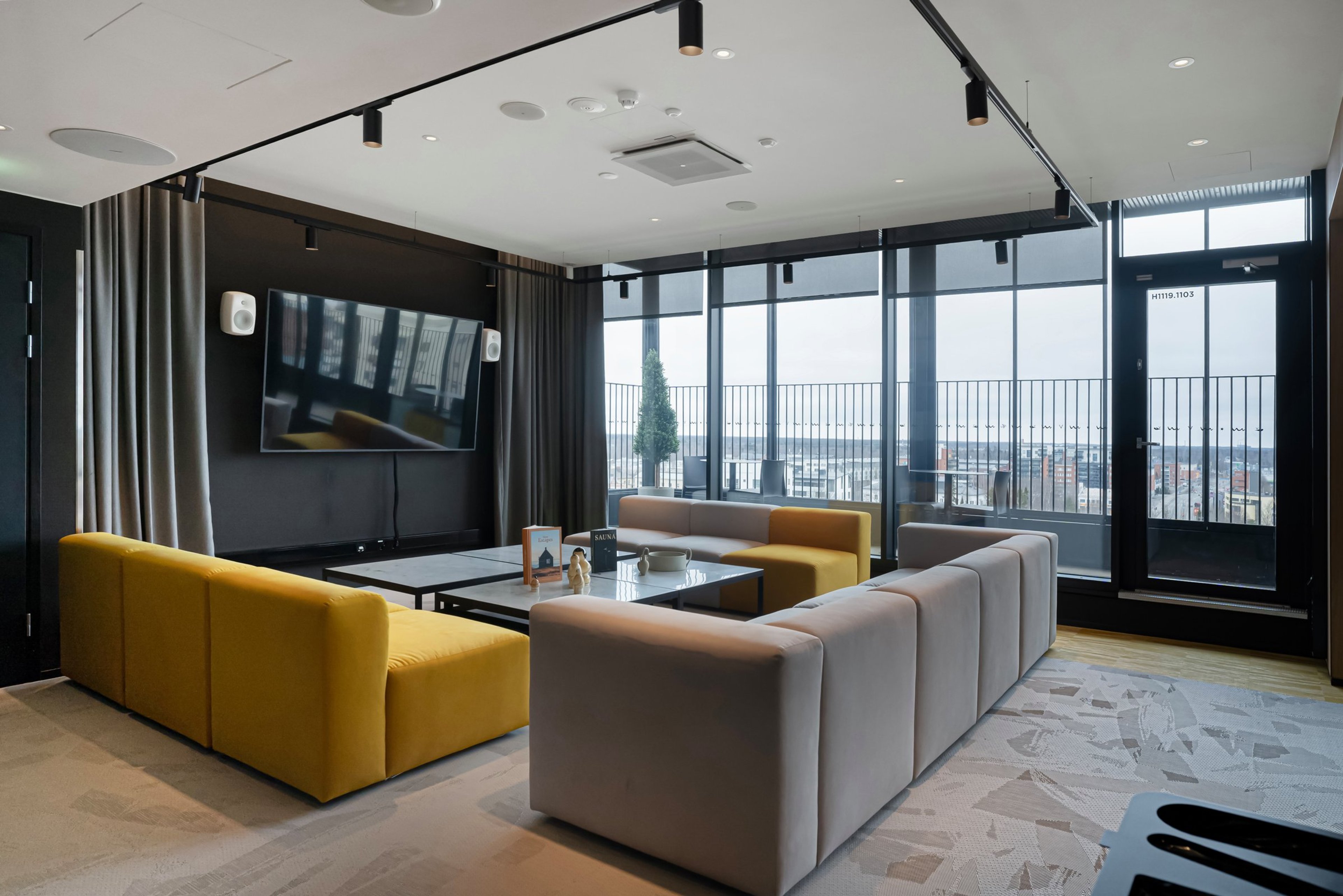 Moderni loungealue, jossa keltaisia ja vaaleita sohvia, iso televisio seinällä ja lattiasta kattoon ulottuvat ikkunat kaupunkinäkymällä.