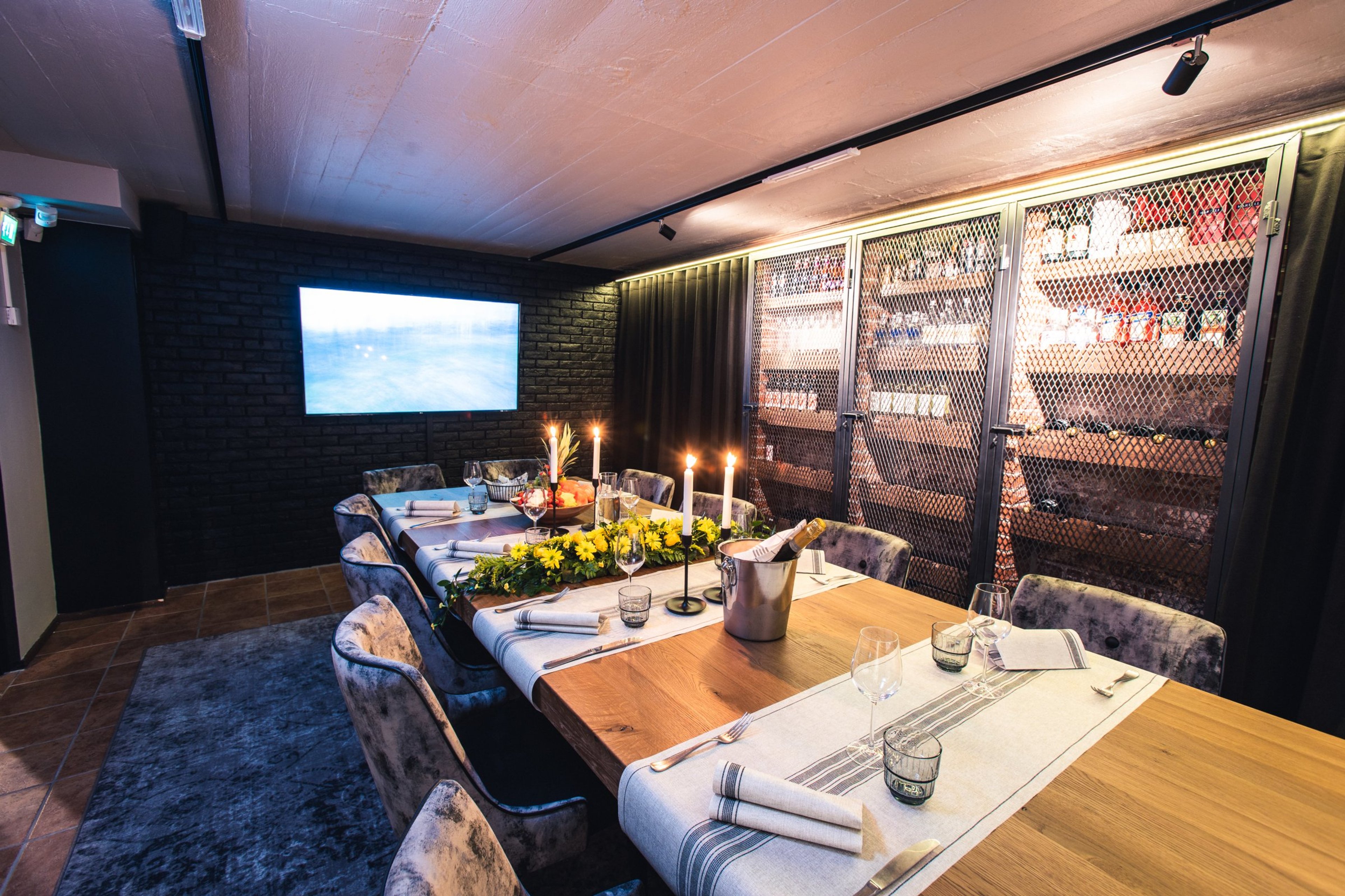 Tunnelmallinen yksityinen ruokailutila, jossa puinen pöytä, samettituolit, kynttilät ja kukkia. Taustalla tiiliseinä ja näyttö.