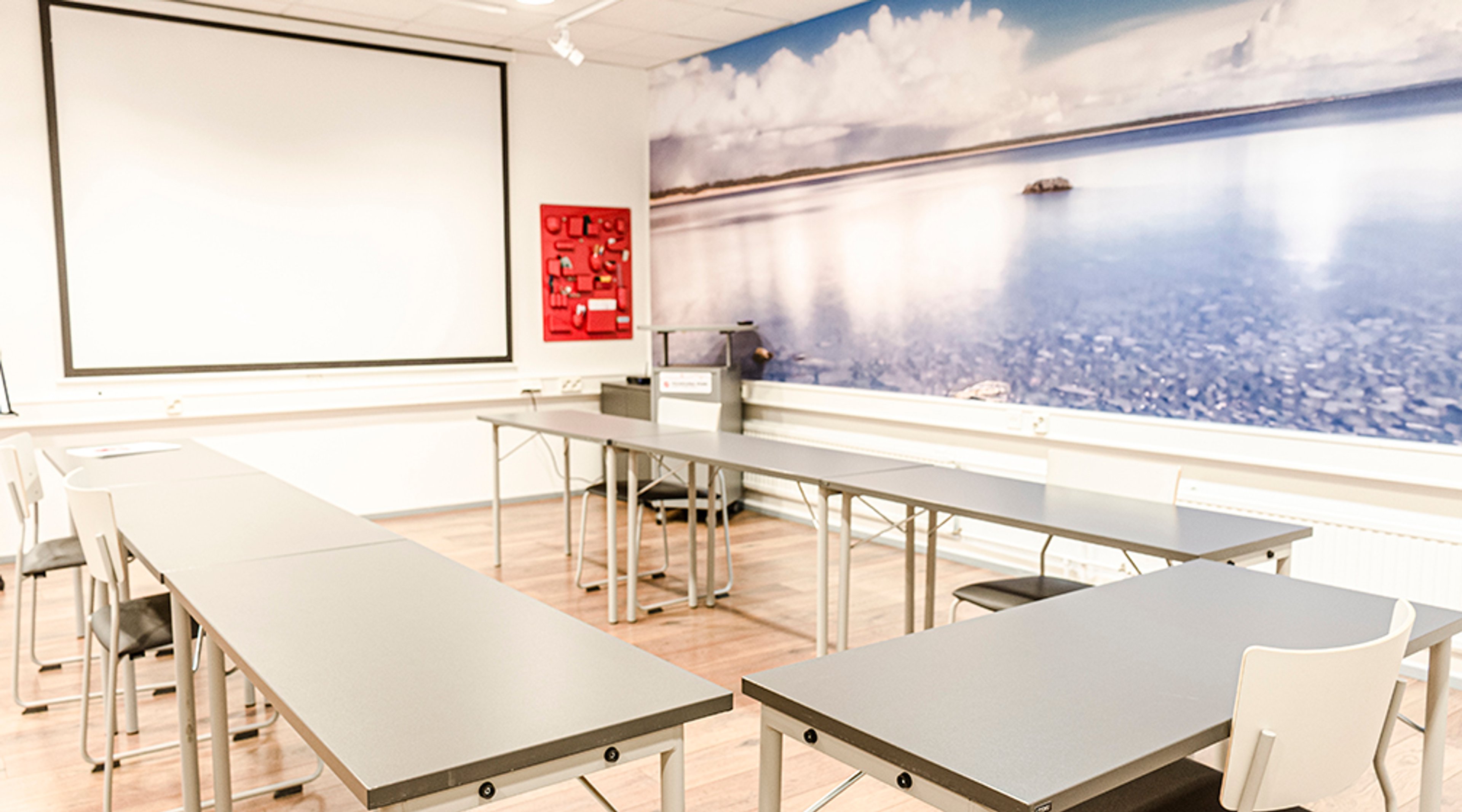 Neuvotteluhuone jossa pöytiä U-muodossa, valkoinen projektorikangas ja seinällä maisematapetti, jossa järvinäkymä.