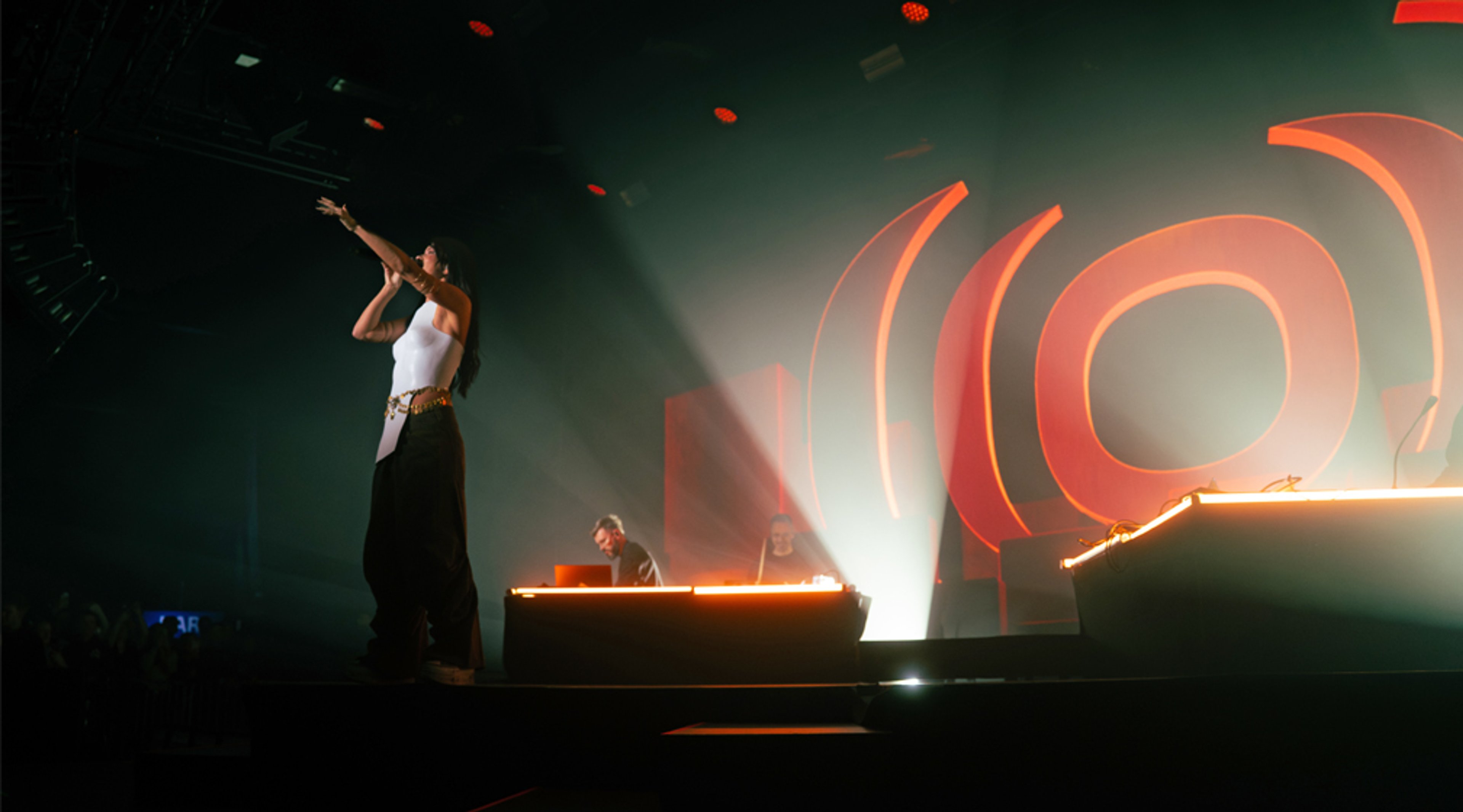 Esiintyjä valkoisessa yläosassa ja mustissa housuissa lavalla, taustalla DJ-pöytä ja oranssi spiraalimainen valoinstallaatio.