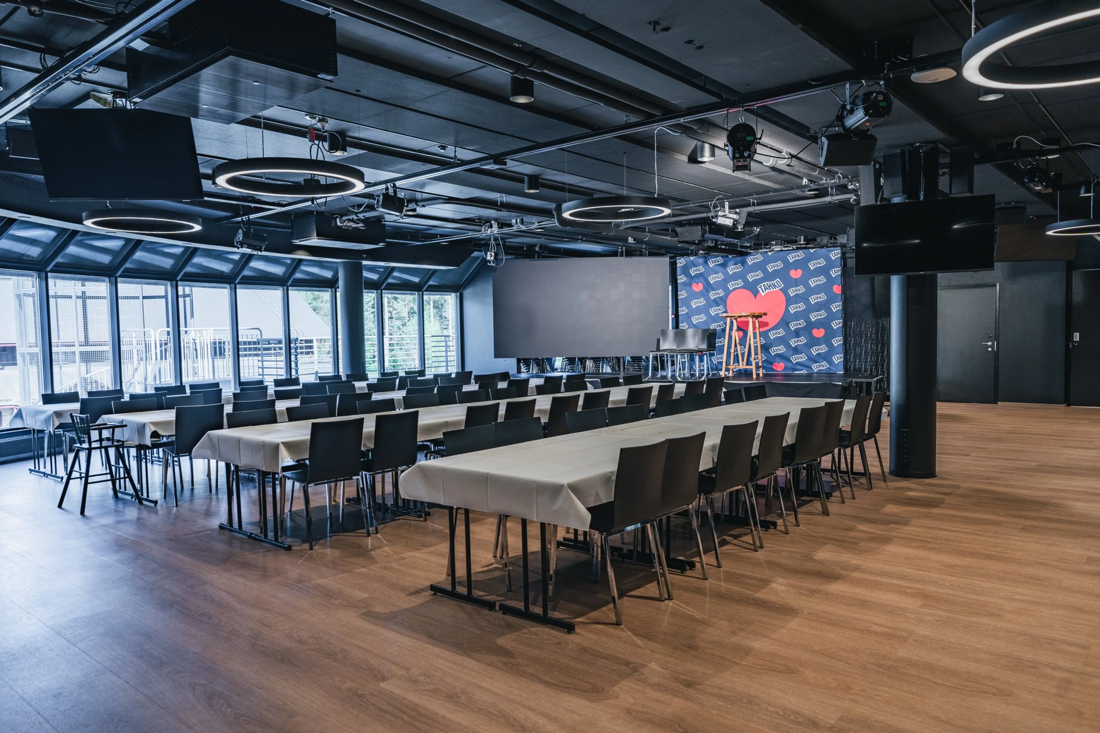 Moderni konferenssihuone, jossa pöydät tuoleineen, lavan edessä näyttö ja taustaseinä. Puulattia, isot ikkunat ja rengasmaiset valaisimet katossa.
