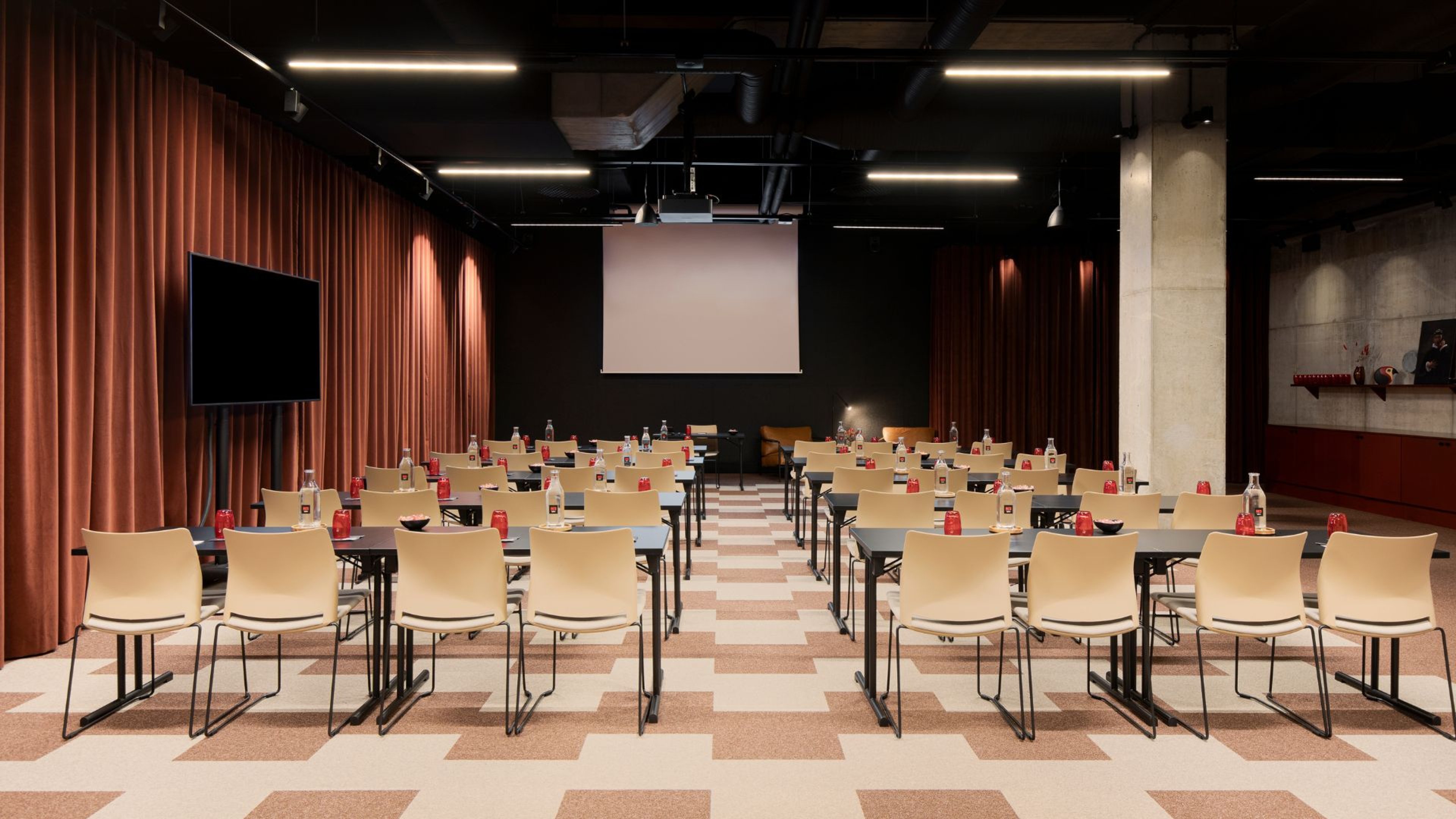 Moderni auditorio ruskeilla verhoilla, riveittäin aseteltuja vaaleita tuoleja ja pöytiä, projektorikangas edessä, tasalattia ja ruutukuvioinen lattia.