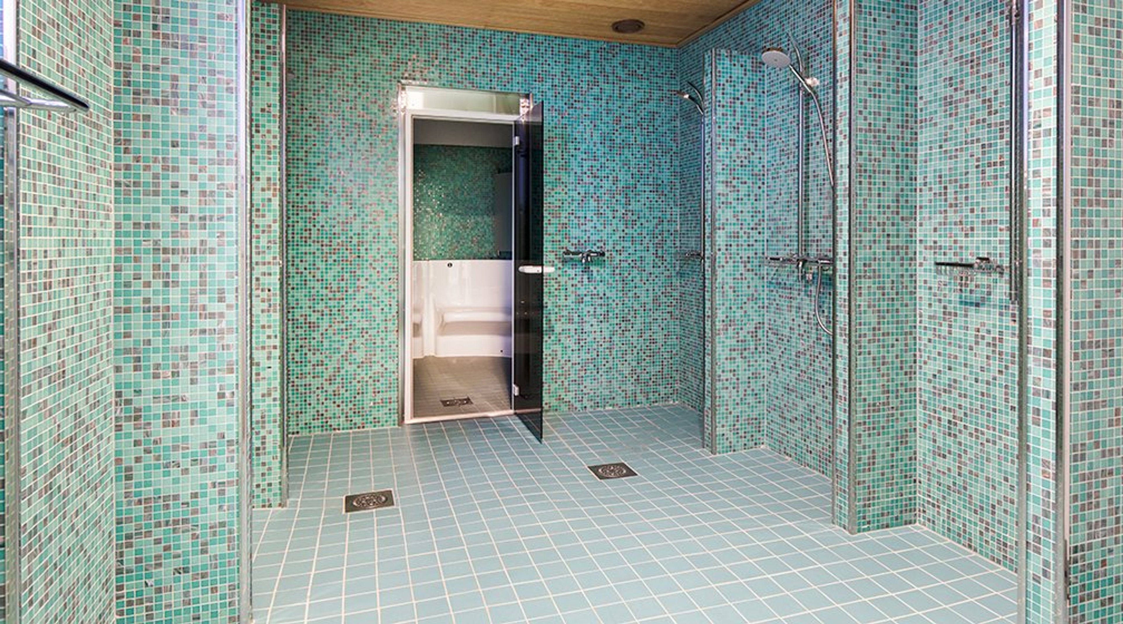 Kylpyhuone turkooseilla mosaiikkilaatoilla, useita suihkuja ja ovi toiseen tilaan. Lattia on tasainen laattalattia.