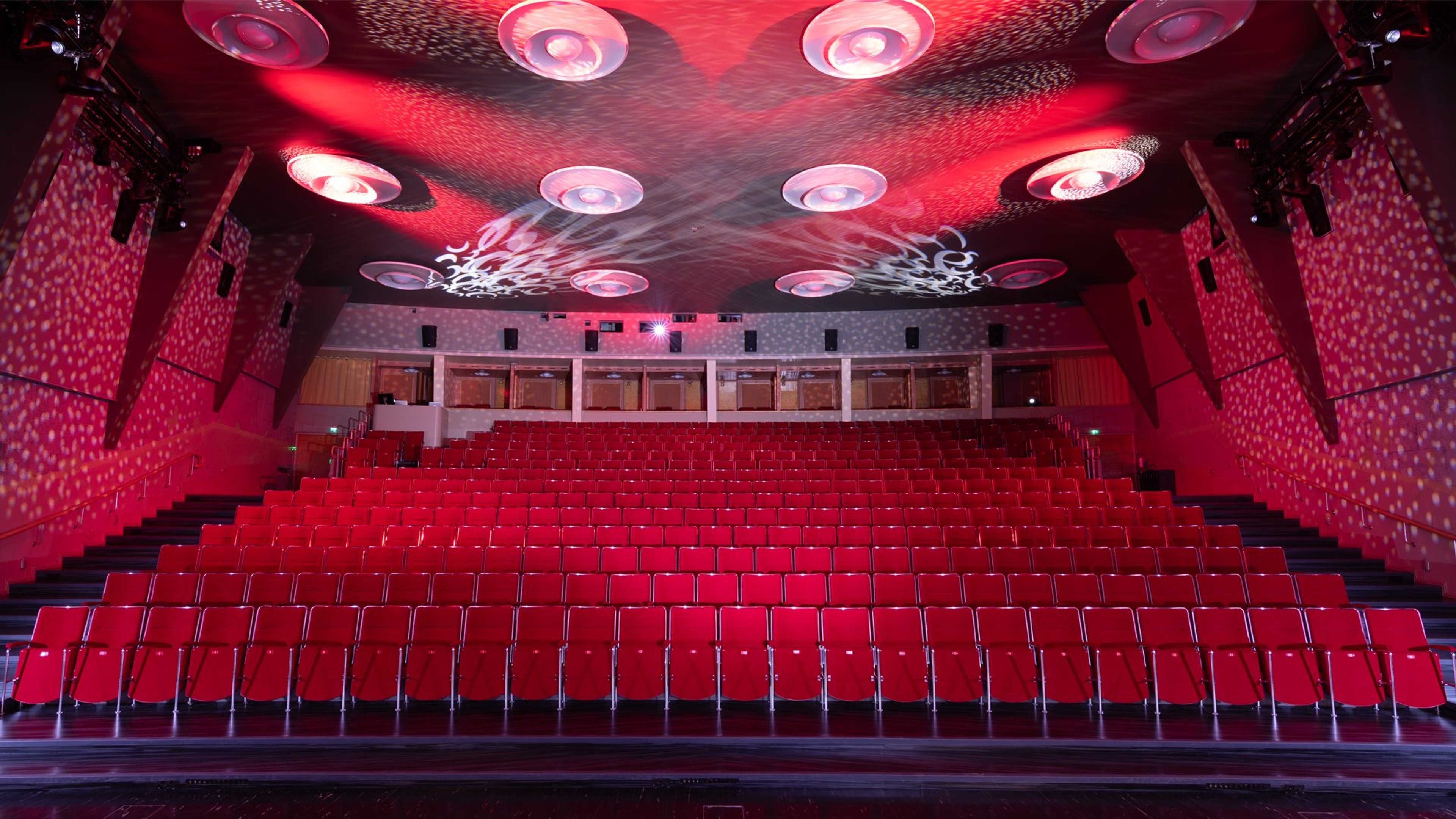 Punaisen auditorion katsomo punaisilla istuimilla, tyhjä sali ja katossa koristeellinen valaistus.