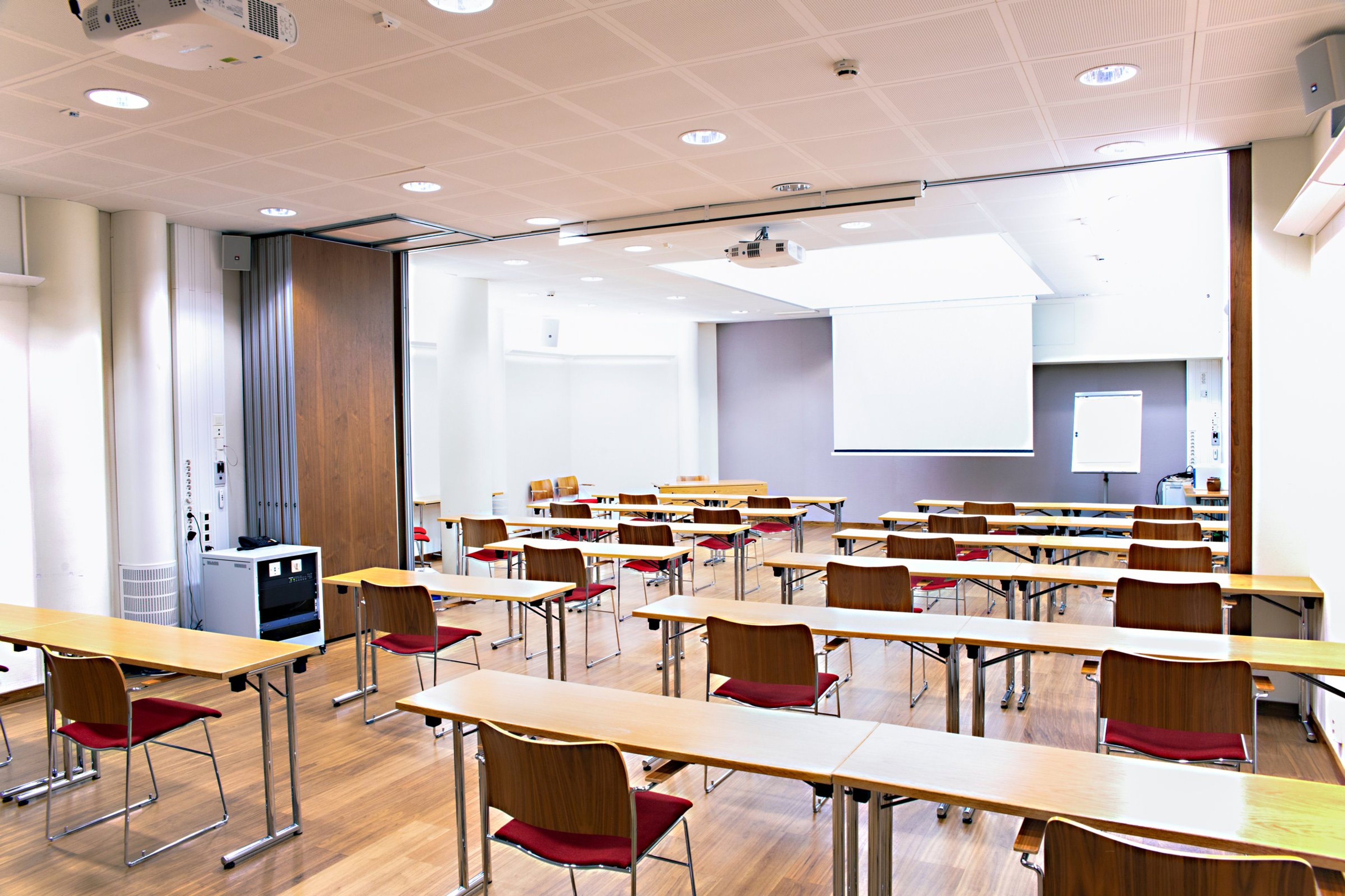 Valoisa luokkahuone puisilla pöydillä ja punaisilla tuoleilla. Tasainen puulattia, kaksi projektoria ja valkokangas edessä. Avara tila taittoseinällä.