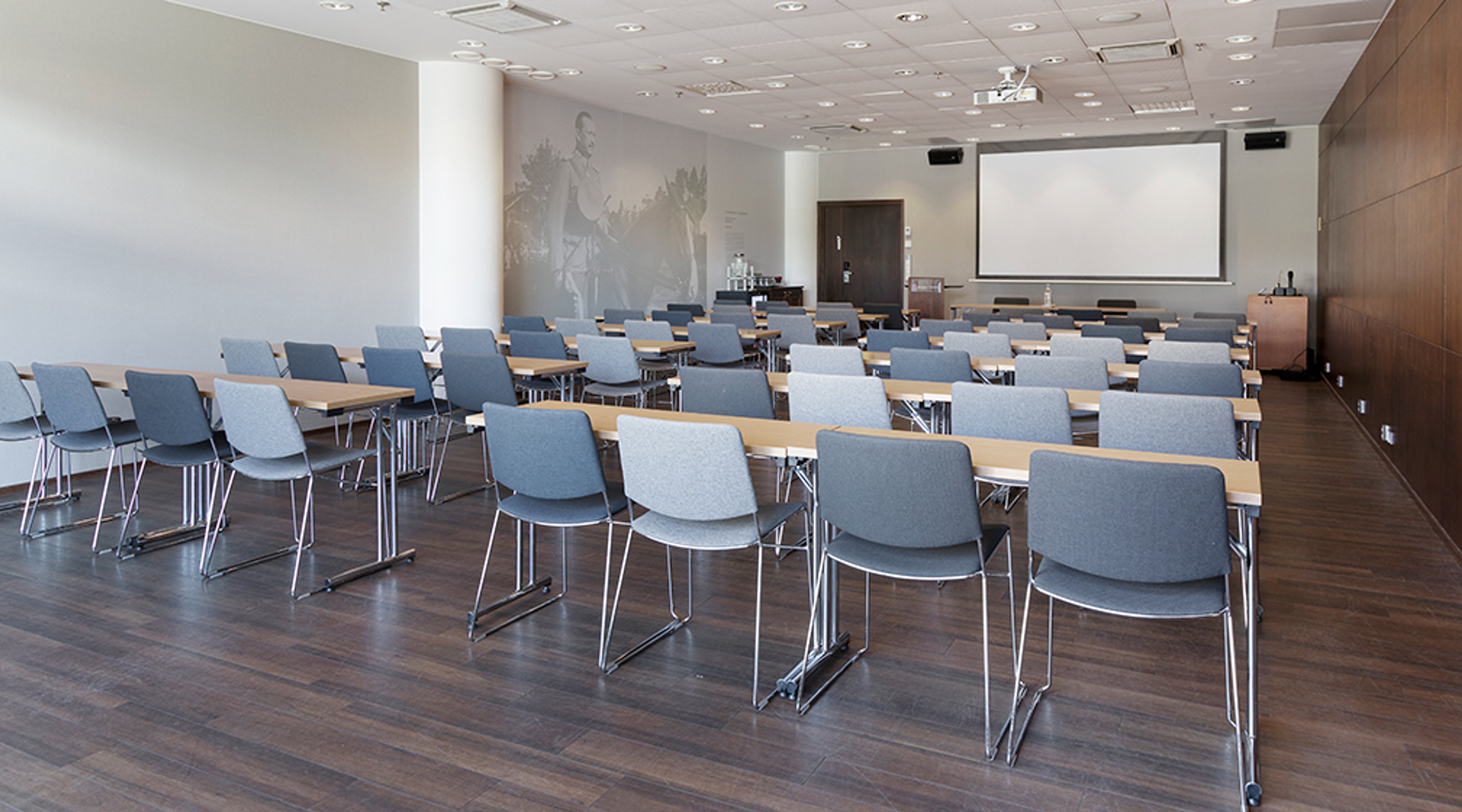 Moderni kokoustila sinisillä tuoleilla ja puisilla pöydillä, valkokangas edessä. Tila on valoisa ja lattialla on tumma puulattia.