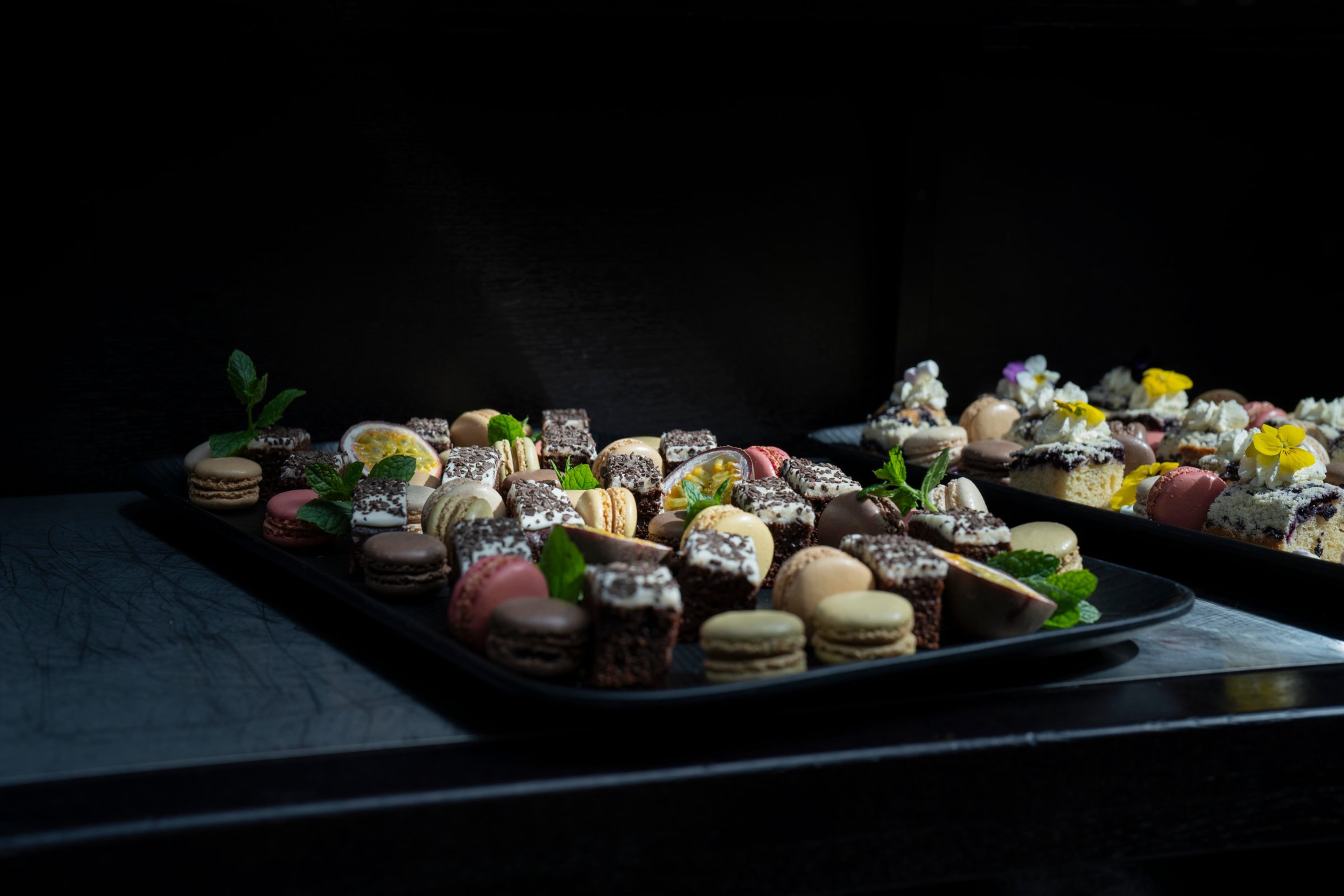 Tarjottimella värikkäitä macaroneja, suklaabrownies-paloja ja muita pieniä leivonnaisia, koristeltu mintunlehdillä ja syötävillä kukilla.