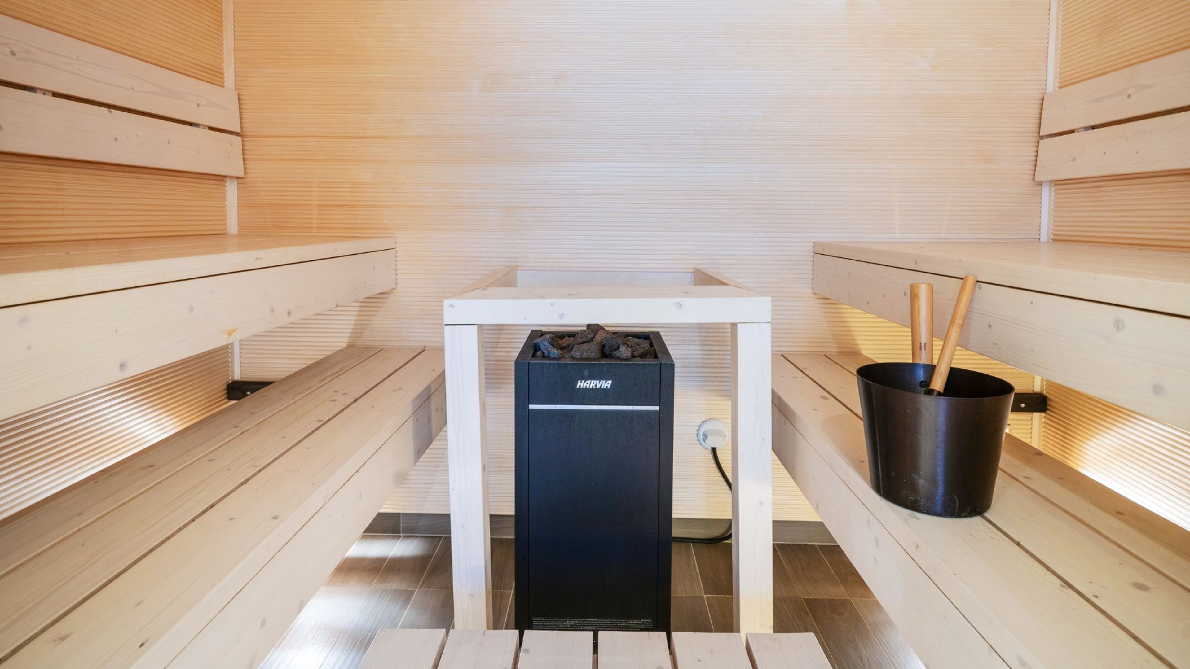 Moderni sauna vaaleilla puupaneeleilla ja penkeillä. Keskellä musta Harvia-kiuas. Lauteiden vieressä musta ämpäri ja löylykauhat.