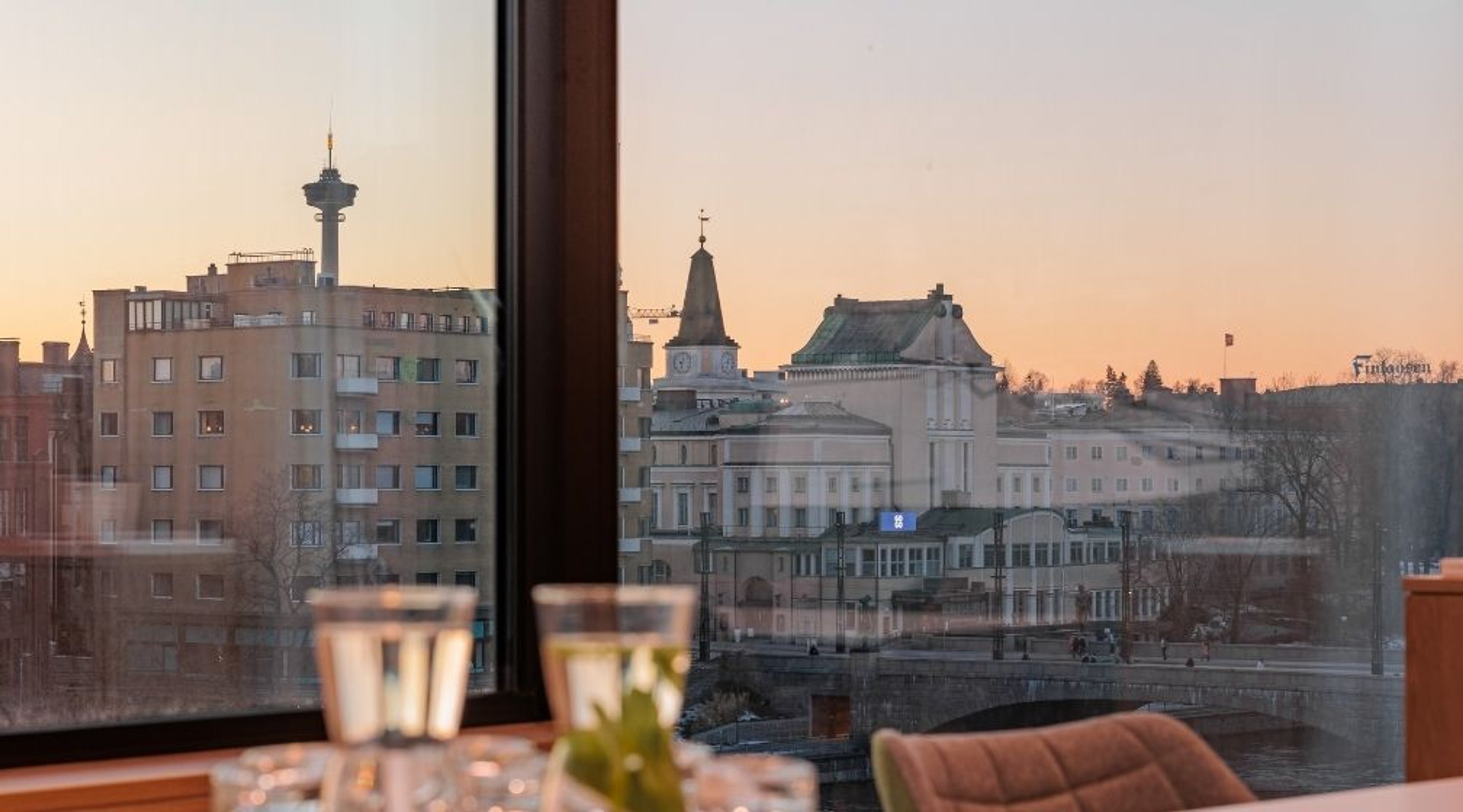 Näkymä ikkunasta Tampereen keskustaan auringonlaskun aikaan. Nähtävissä Näsinneula-torni, kellotorni ja historiallisia rakennuksia joen rannalla.