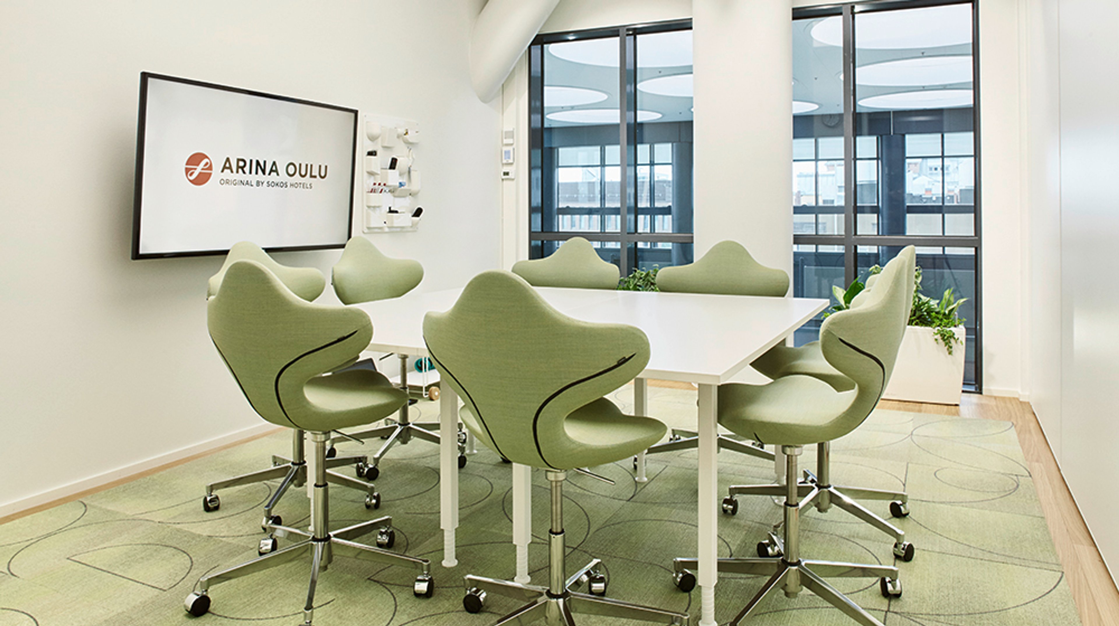 Moderni neuvotteluhuone vihreillä ergonomisilla tuoleilla ja valkoisella pöydällä. Vaalea sisustus, vihreä matto ja näyttö seinällä.