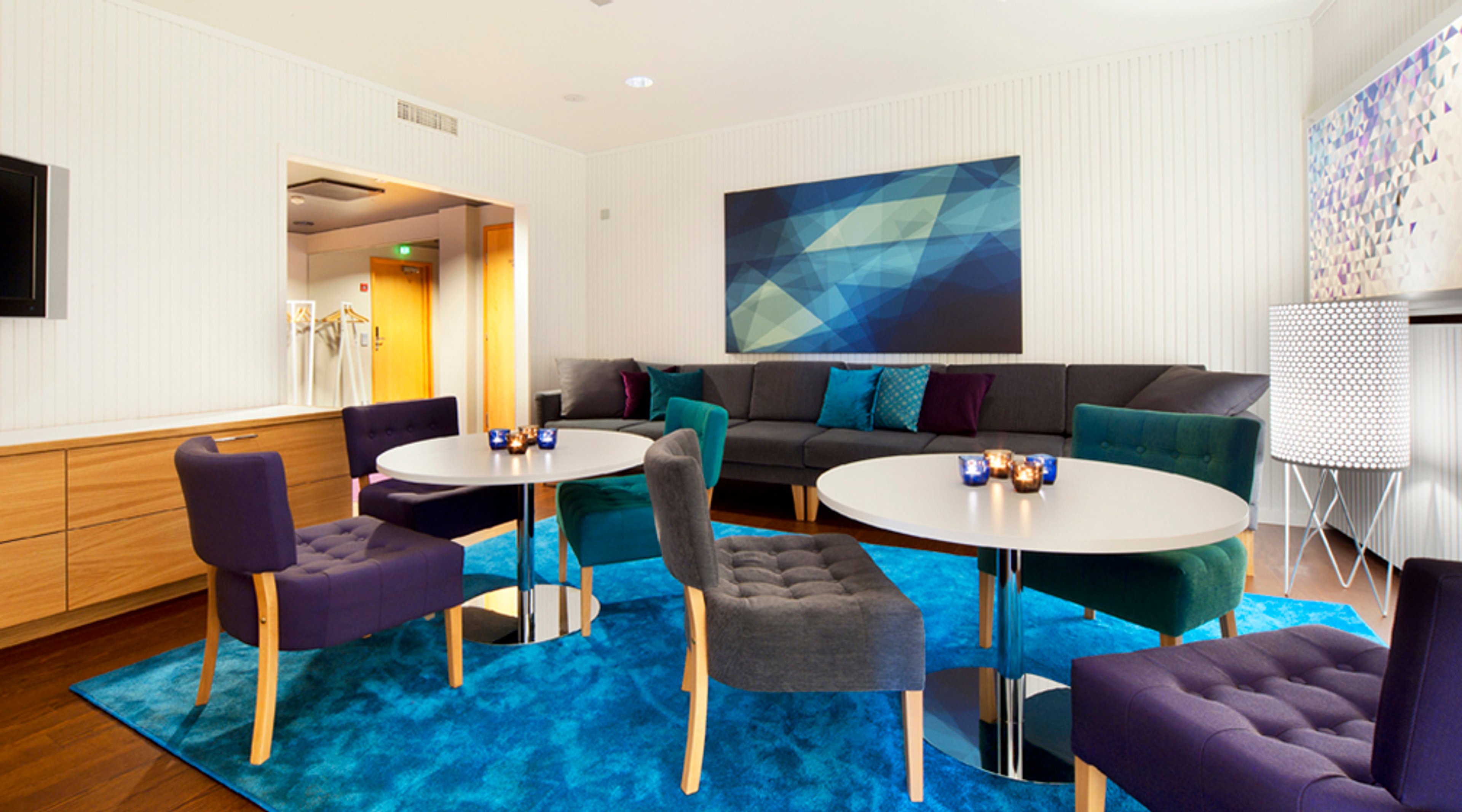 Moderni loungealue, jossa harmaa sohva, pyöreät pöydät ja värikkäät tuolit sinisellä matolla. Seinällä sinisävyinen taide.