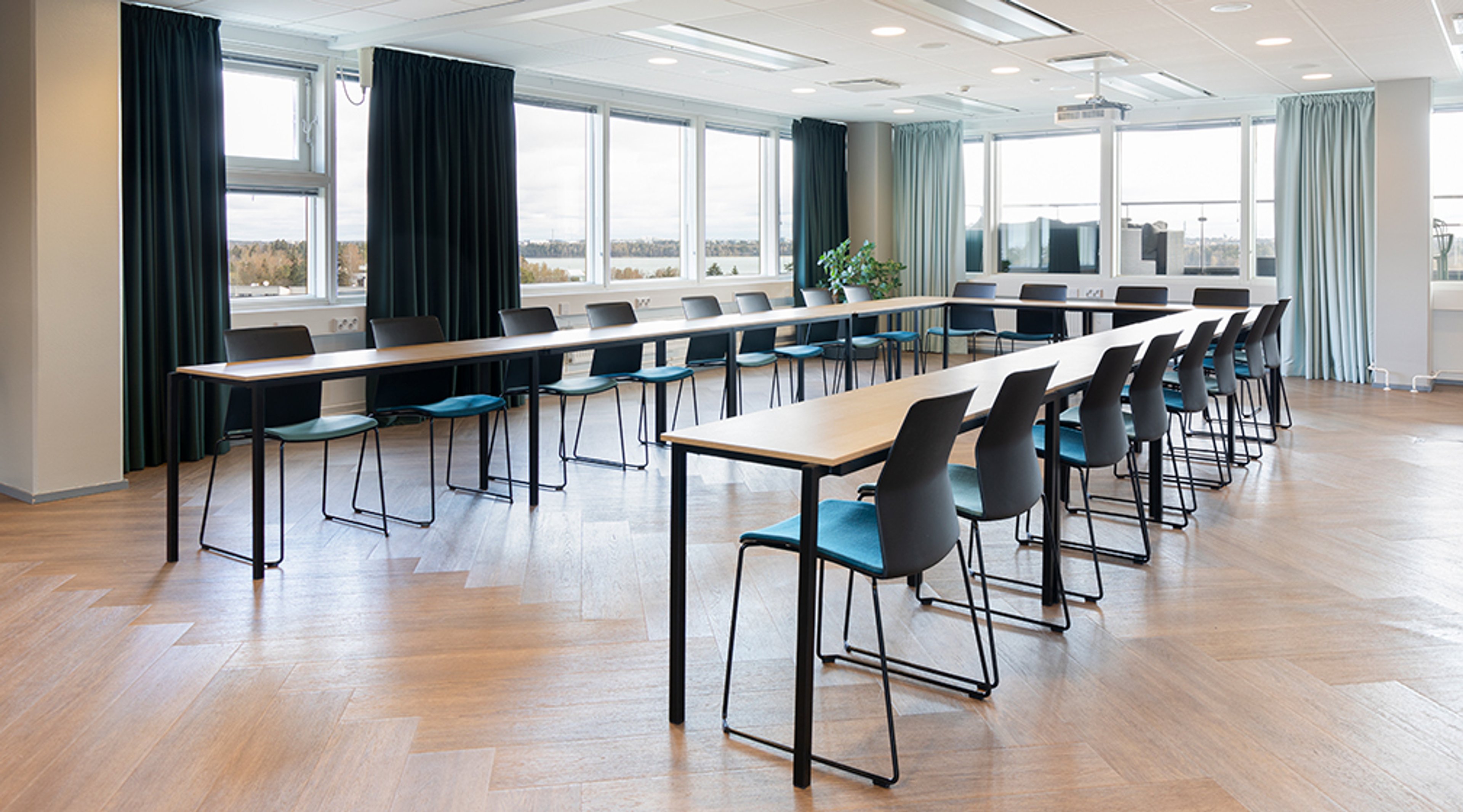Modern meeting room with long tables and black-blue chairs, large windows with landscape view. Flat wooden floor and good lighting.