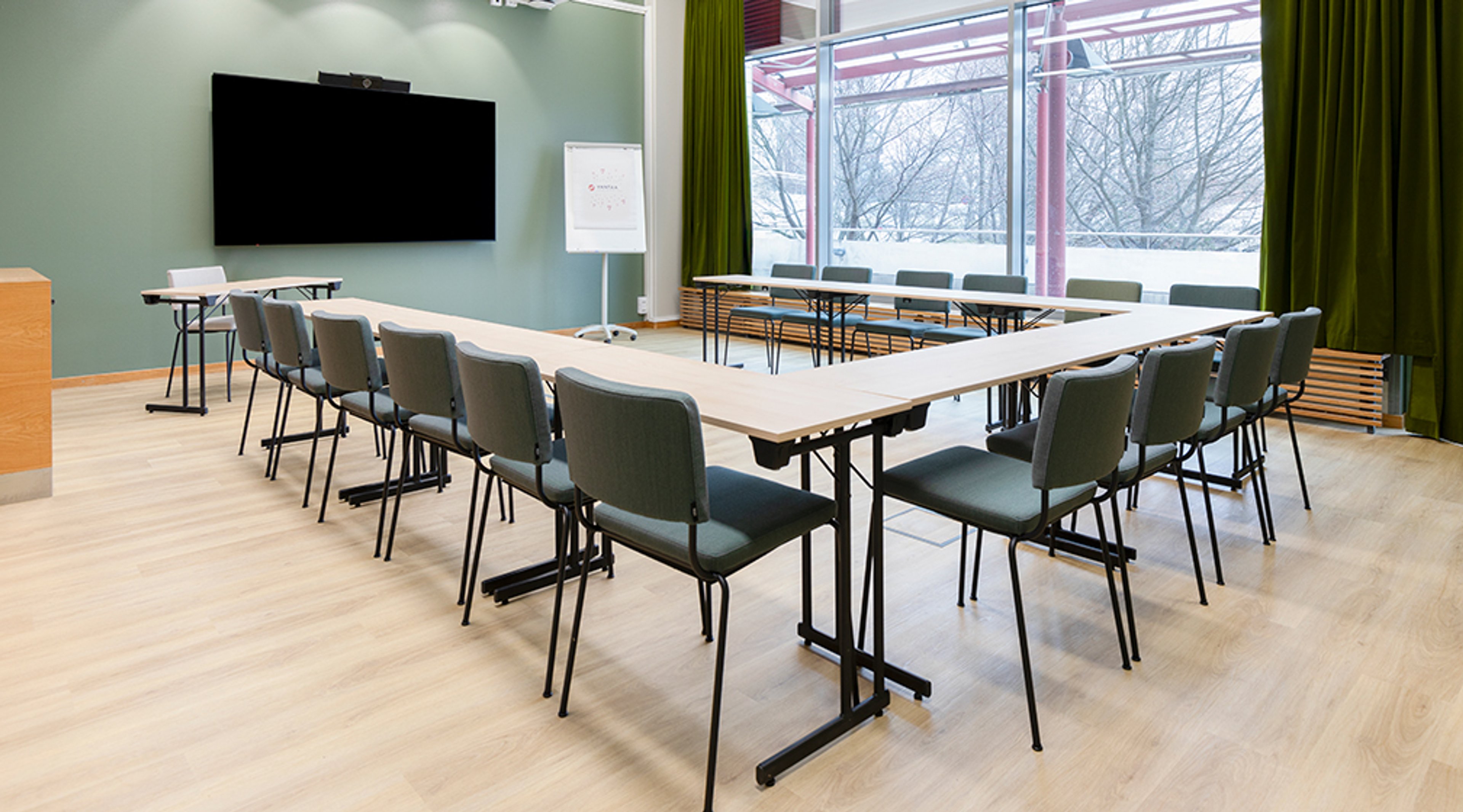 Valoisa kokoushuone U-muotoisella pöytäjärjestelyllä, seinällä iso näyttö ja taustalla suuret ikkunat talviseen maisemaan.