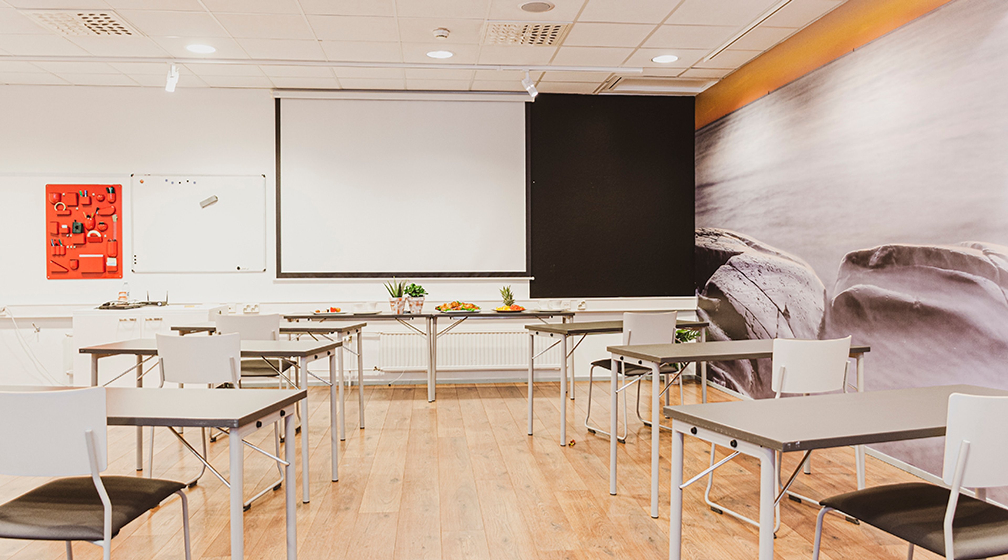 Valoisa kokoushuone puulattialla, jossa valkoisia pöytiä ja tuoleja. Edessä valkokangasvalkokangas, valkotaulu ja koristeellinen seinäkuvitus.