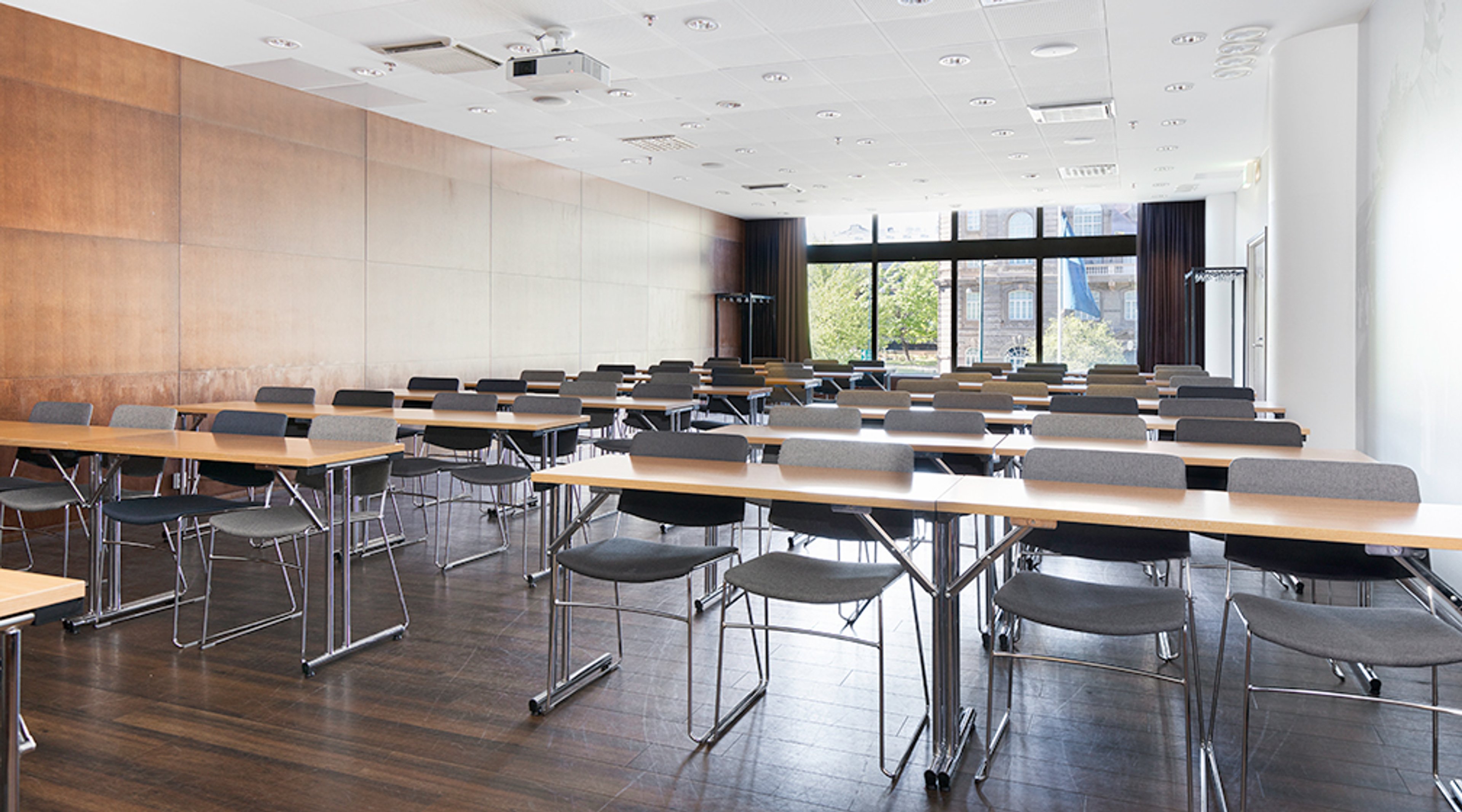 Moderni kokoustila, jossa on puiset pöydät ja harmaat tuolit rivistöissä, suuret ikkunat ja osittain puupaneeliset seinät. Tilassa on kova lattia.