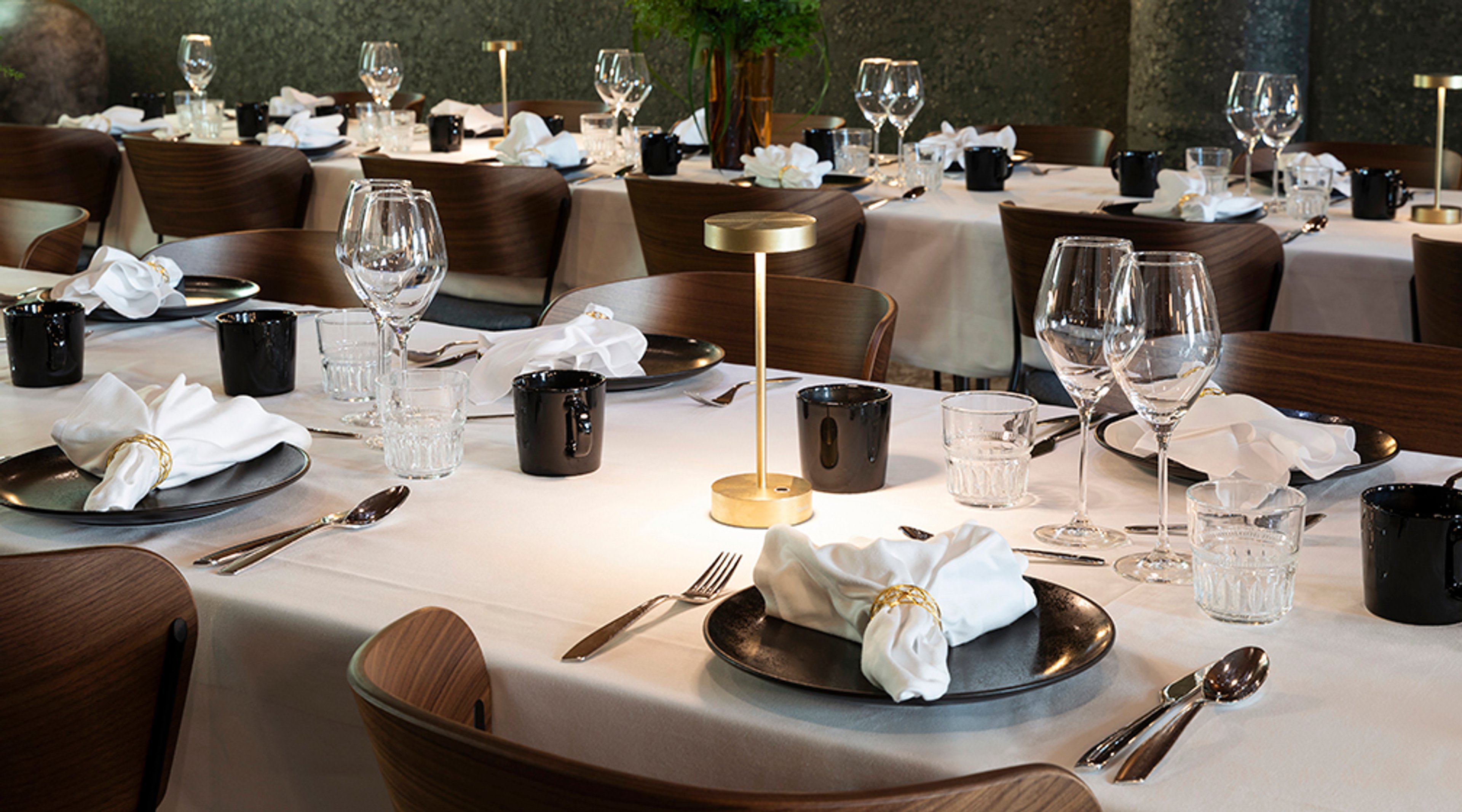 Tyylikkäästi katetut ravintolan pöydät valkoisilla liinoilla, tummilla lautasilla ja kultaisilla valaisimilla. Puiset tuolit kehystävät tilaa.