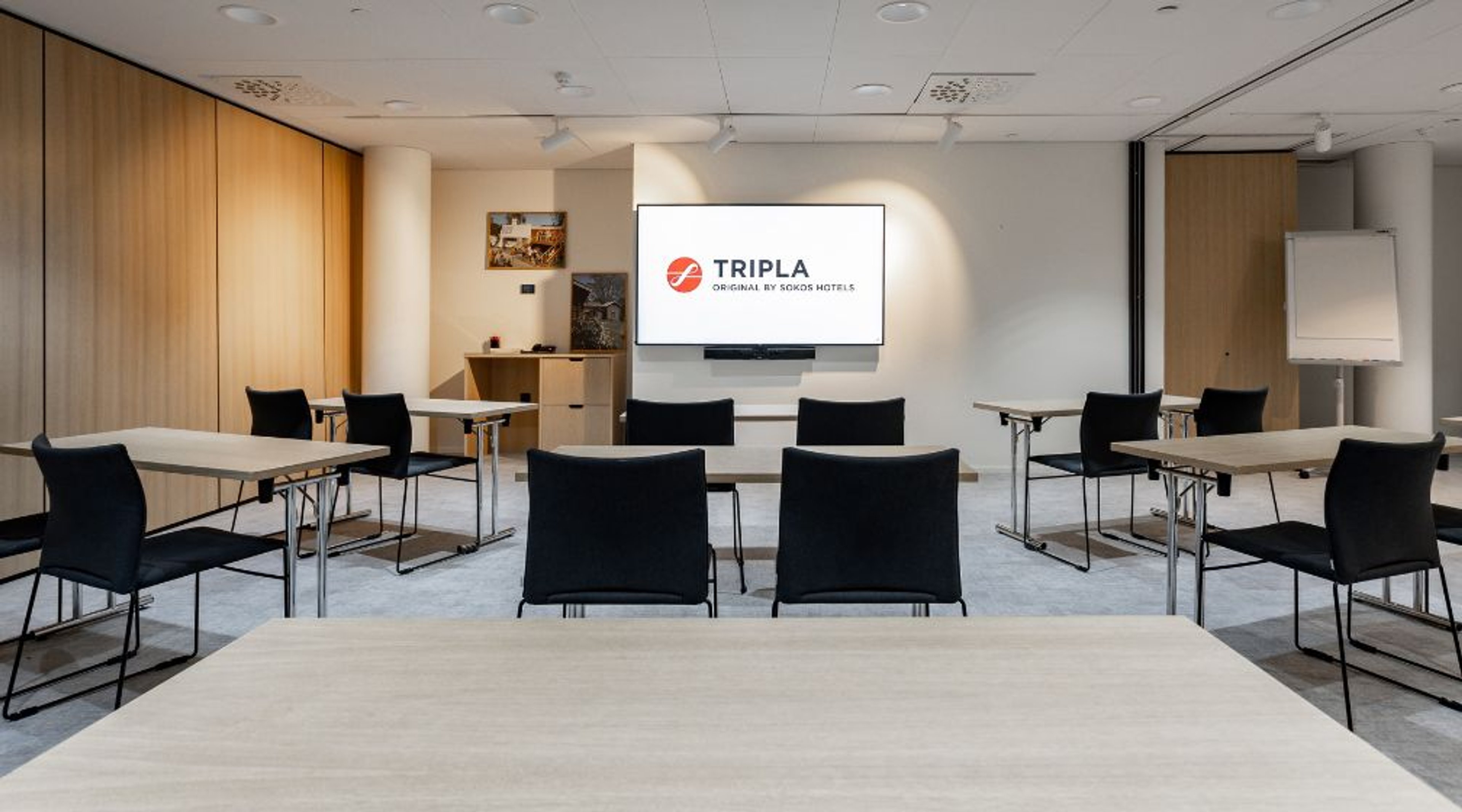 Moderni kokoustila, jossa vaaleita puisia pöytiä ja mustia tuoleja. Valkoisella seinällä suuri näyttö. Tasainen lattia ja hyvä valaistus.