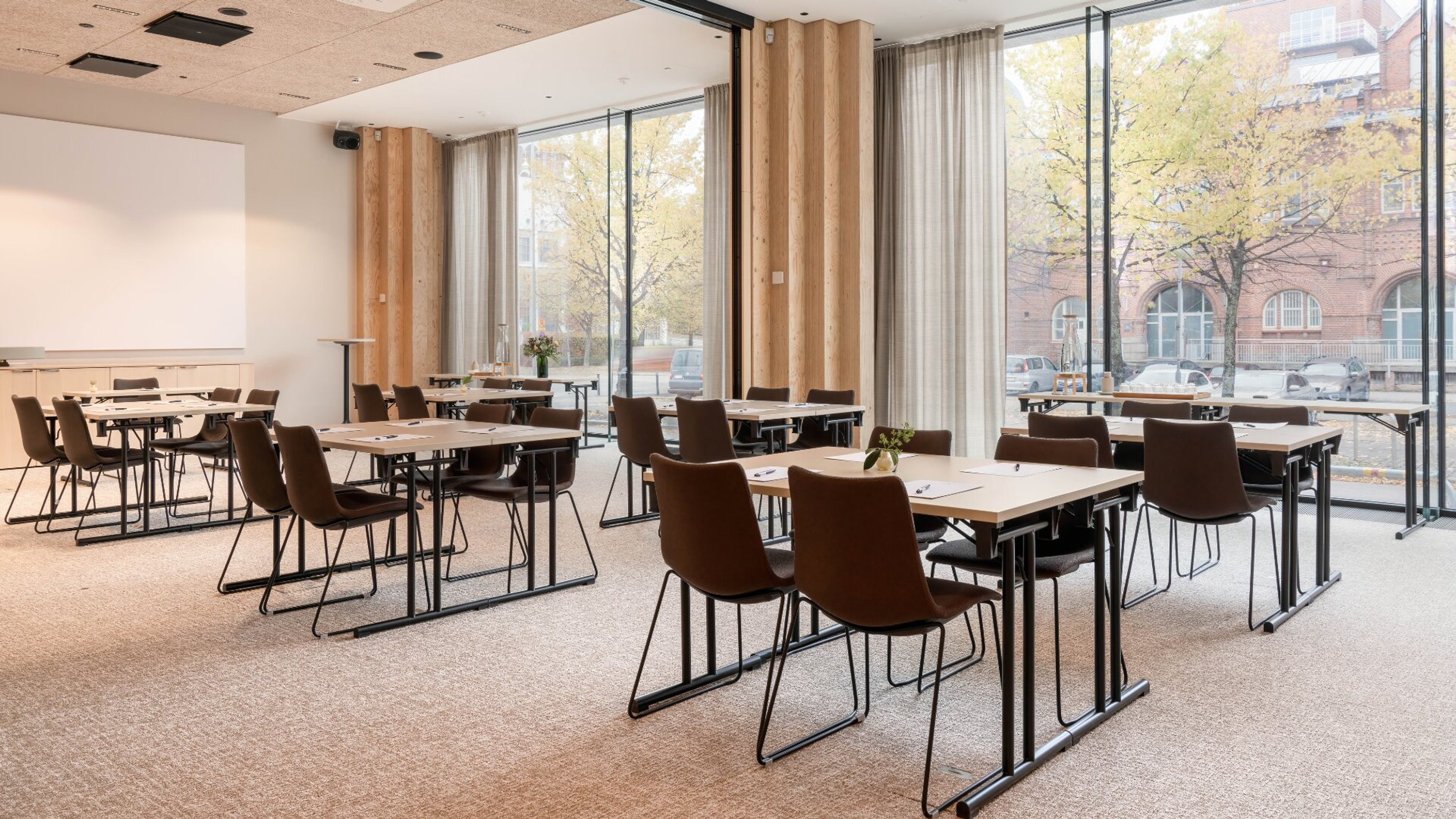 Moderni kokoushuone, jossa vaaleita puupöytiä ja ruskeita tuoleja. Lattialla matto ja seinillä suuret ikkunat, joista näkyy puita ja tiilirakennus.