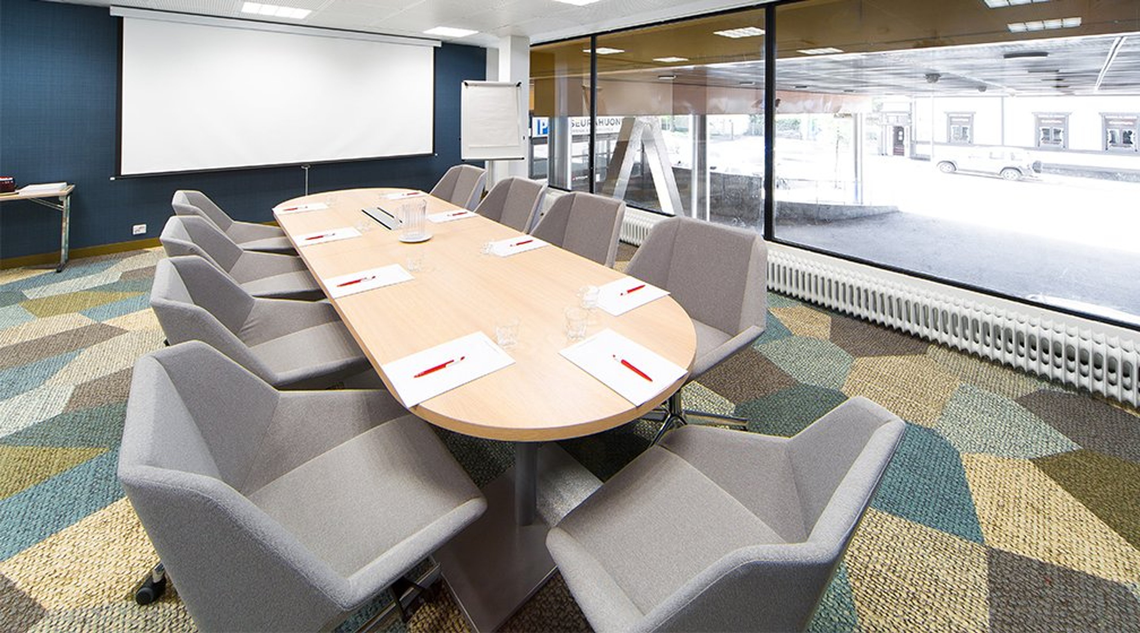 Kokoushuone, jossa pitkä puinen pöytä harmain tuolein, valkokangas seinällä ja suuret ikkunat. Lattia on värikäs kuviollinen kokolattiamatto.