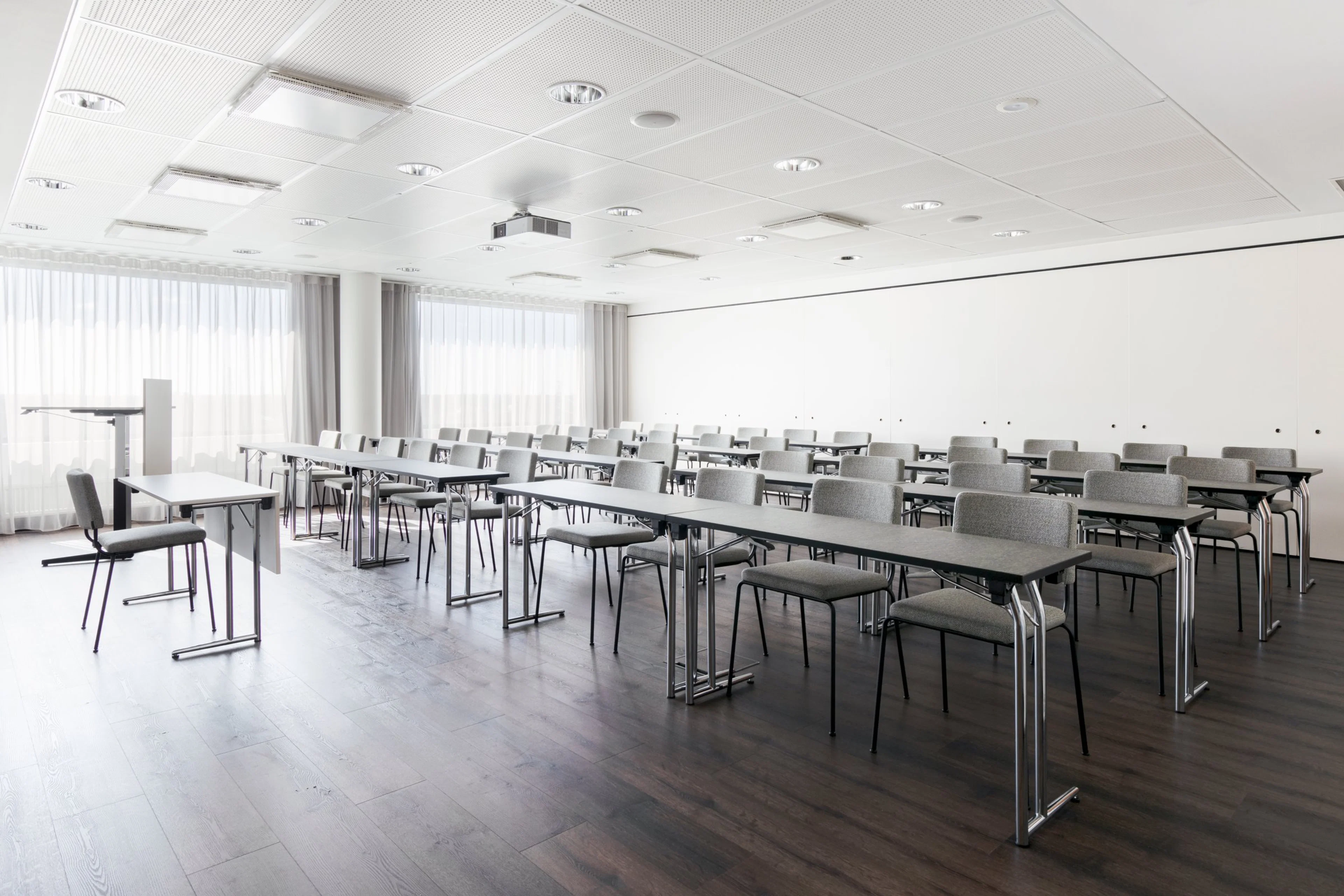 Moderni, vaalea kokoustila, jossa on pitkiä pöytiä ja harmaita tuoleja riveissä. Huoneessa on tasainen puulattia ja hyvä valaistus katossa.