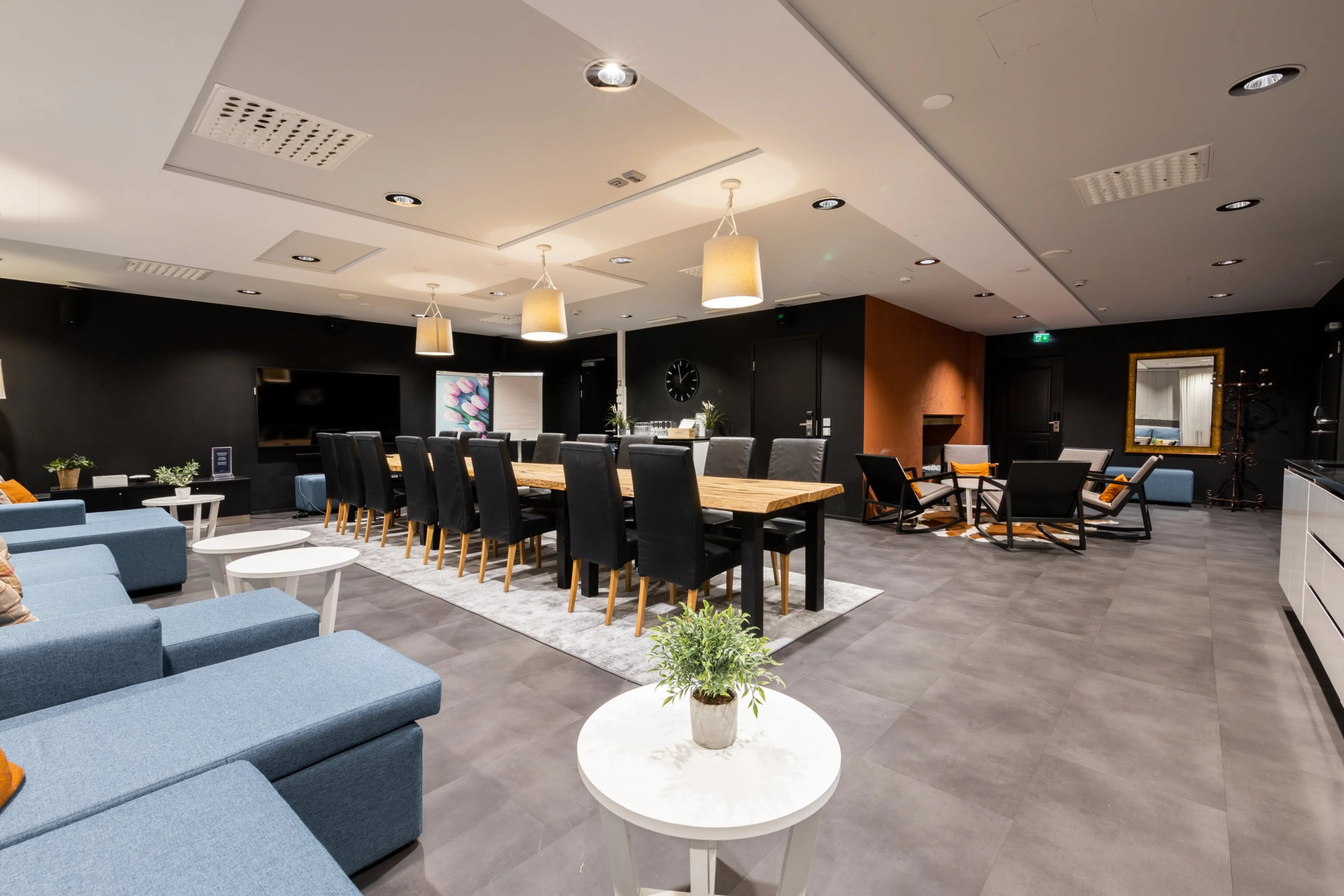 Moderni kokoustila, jossa puinen ruokapöytä mustilla tuoleilla, sinisiä sohvia, valkoisia pöytiä ja tasainen harmaa lattia. Hyvin valaistu.