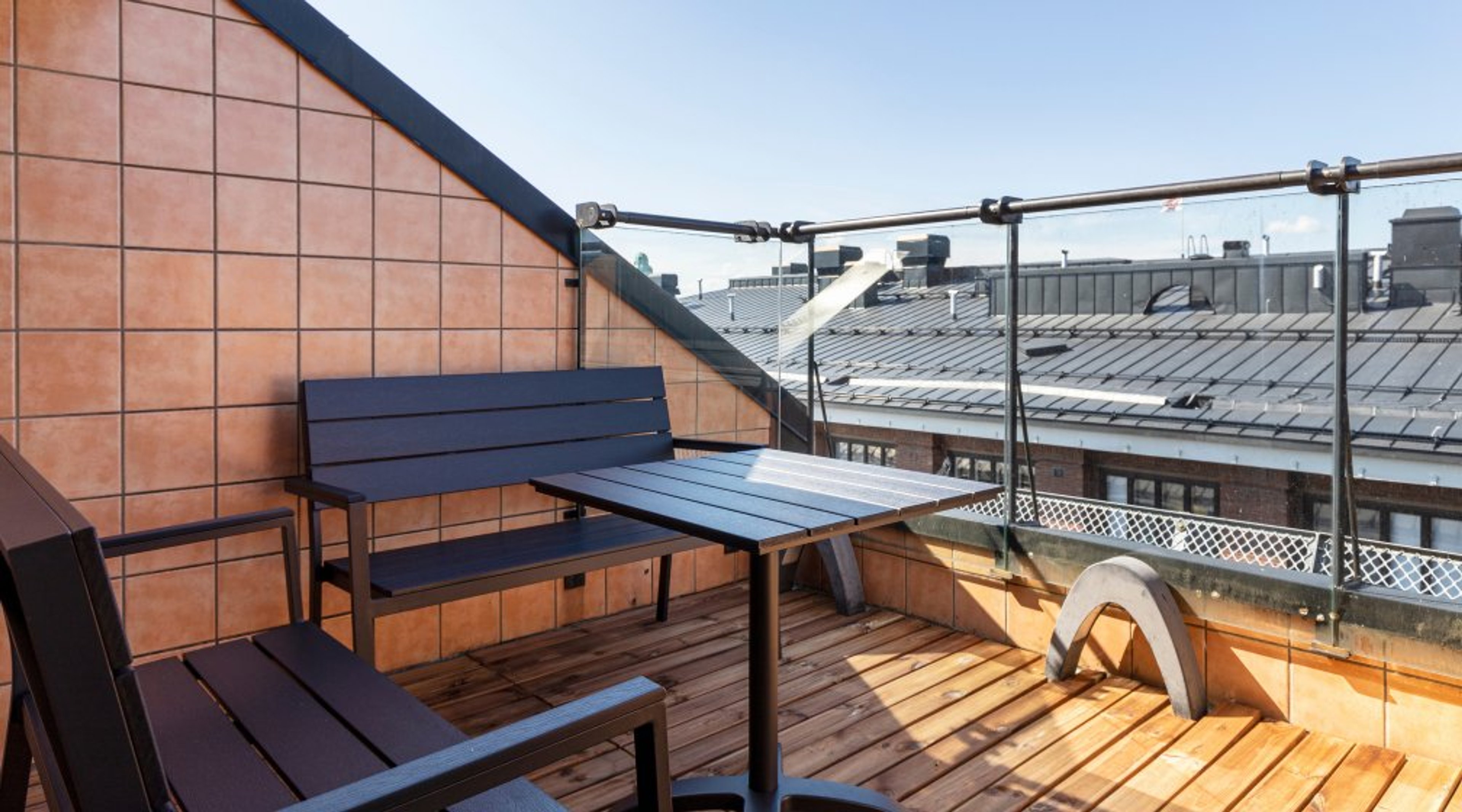 Rooftop terrace with blue bench and table on wooden deck, glass railings, and view of city rooftops.