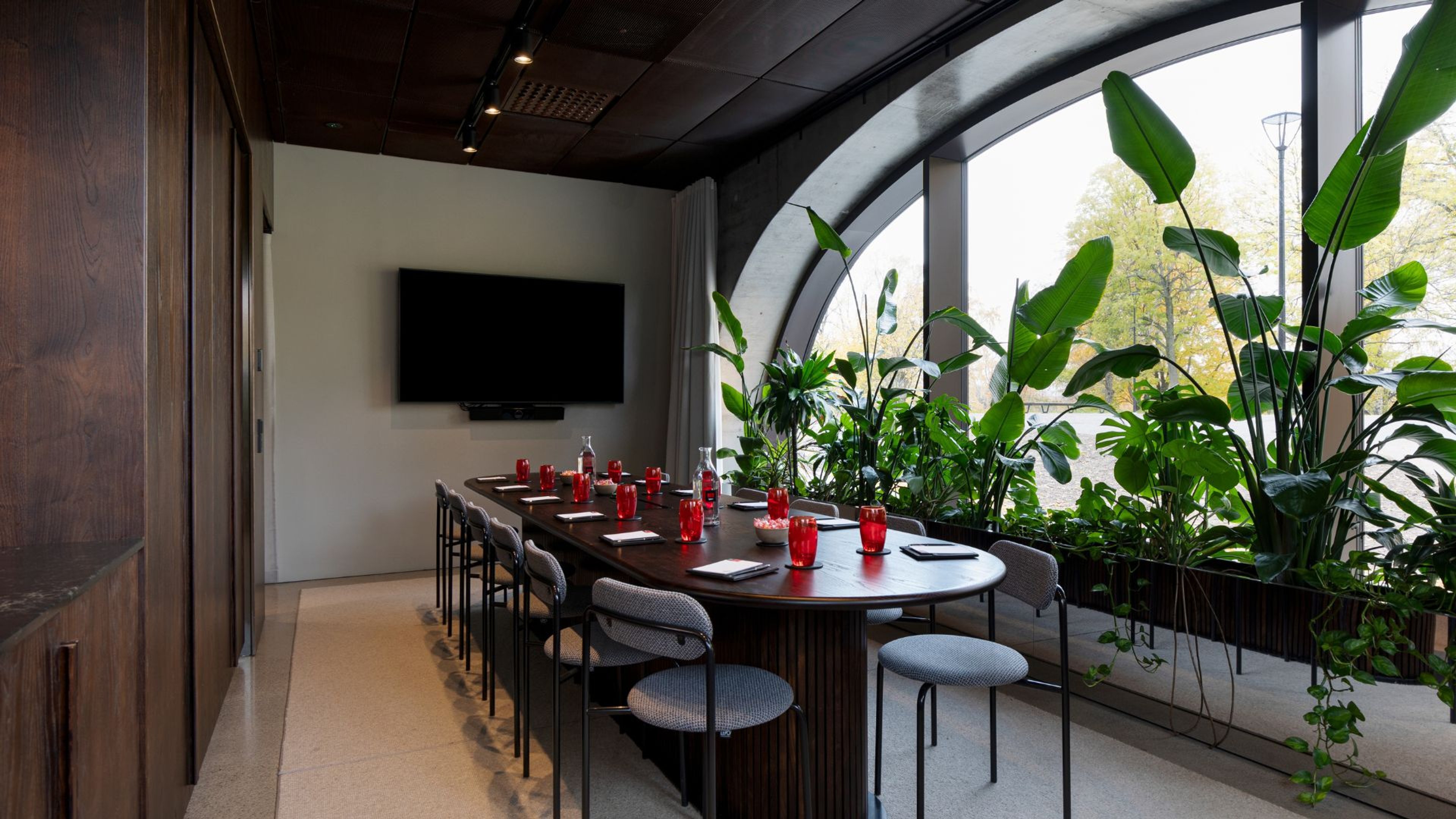 Moderni kokoushuone, jossa puinen pöytä, punaiset juomalasit ja runsaasti viherkasveja kaarevien ikkunoiden edessä. Seinällä televisio.