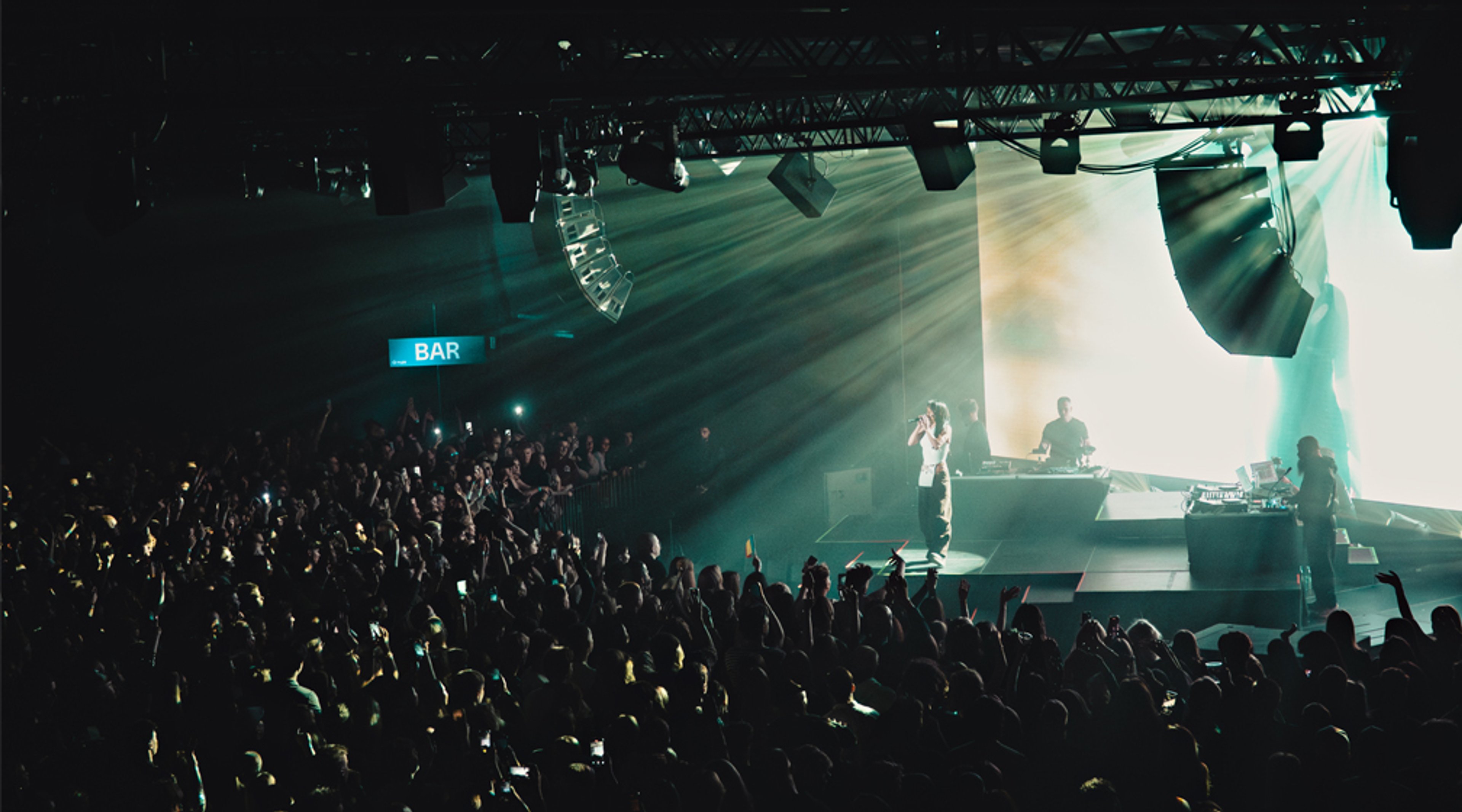 Konserttitila, jossa esiintyjät lavalla ja suuri yleisö. Vihreät valonsäteet leikkaavat hämärää tilaa, taustalla on baari.