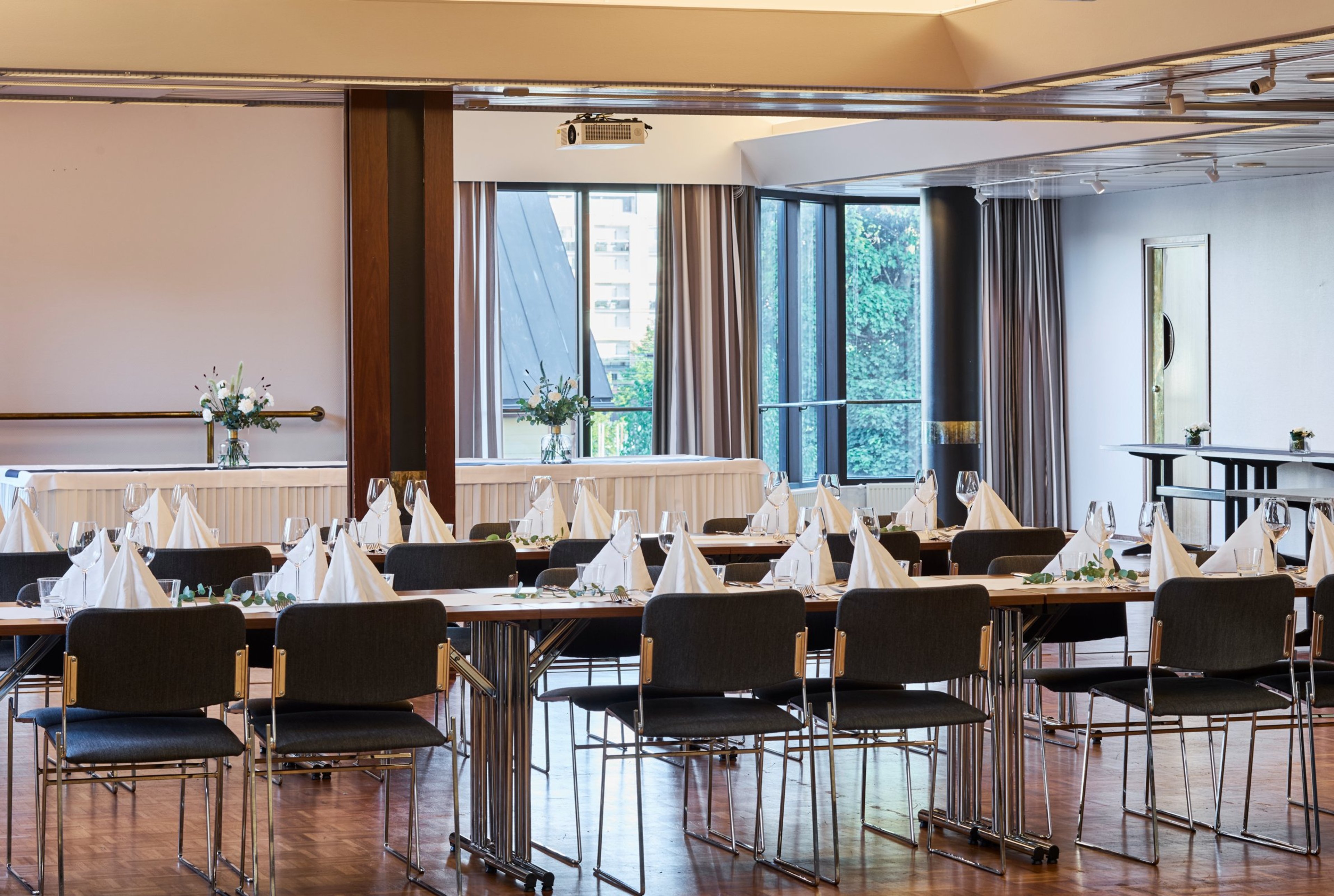 Tilava juhlasali lautasten ja servettien kattauksella. Taustalla suuret ikkunat, puinen lattia ja videotykki katossa. Elegantisti aseteltu pöytäryhmä.