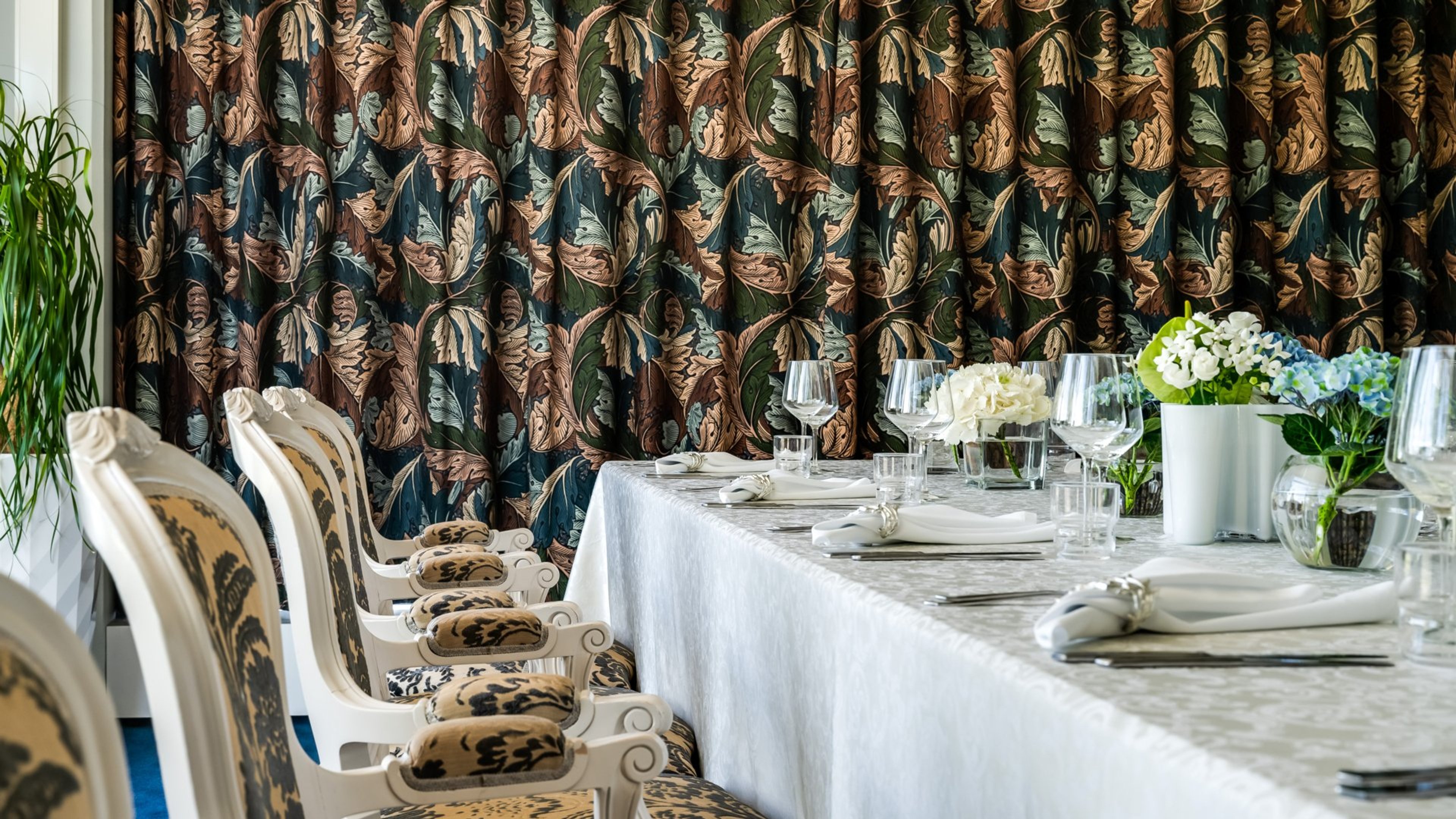 Tyylikäs ravintolan pöytä valkoisella liinalla ja kukka-asetelmilla. Taustalla näyttävät lehtiaiheisella kankaalla päällystetyt seinät ja valkoiset koristeelliset tuolit.