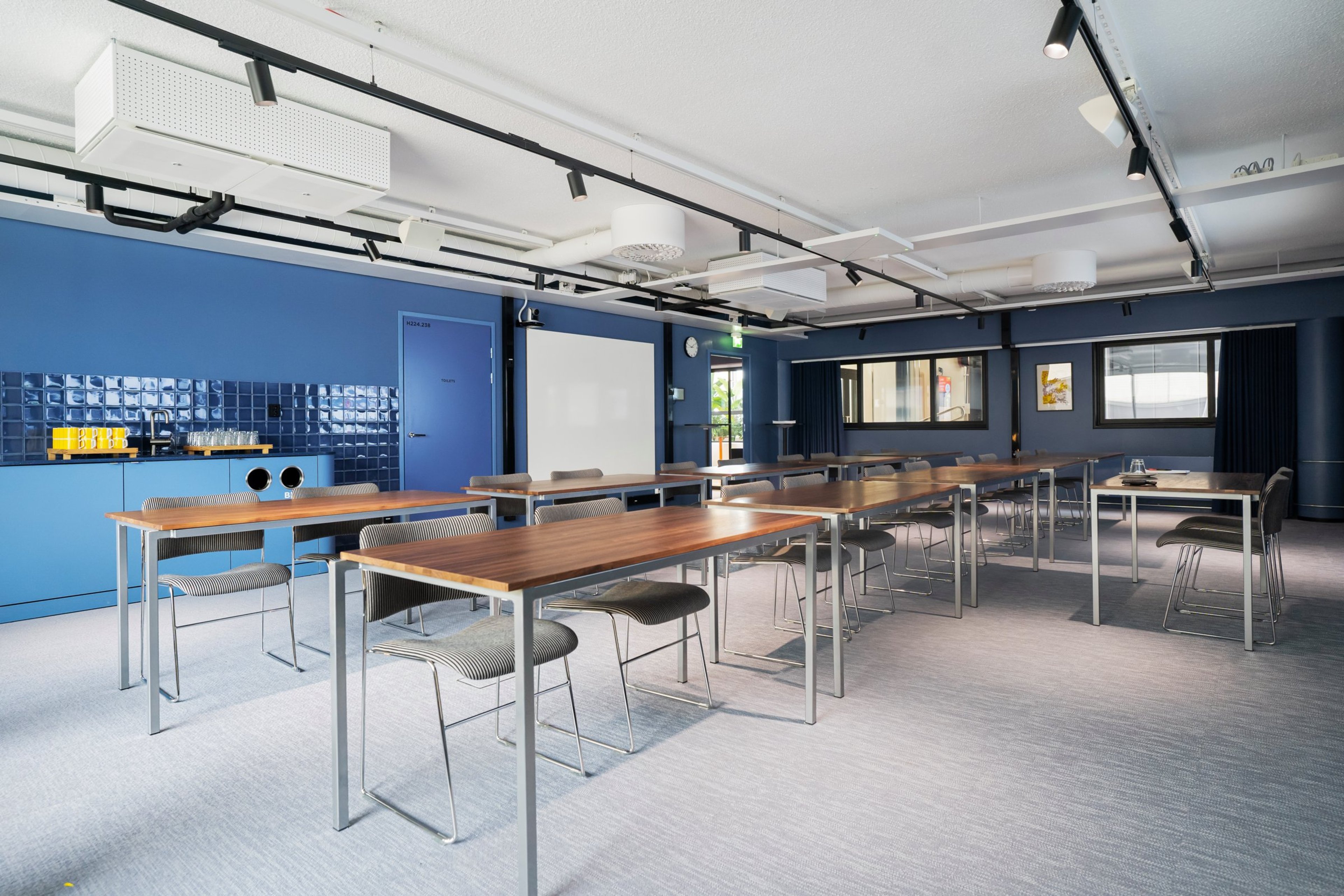 Moderni kokoustila sinisillä seinillä, puupintaisilla pöydillä ja raidallisilla tuoleilla. Taustalla projektori ja tarjoilupiste. Tasainen lattiamatto ja hyvä valaistus.